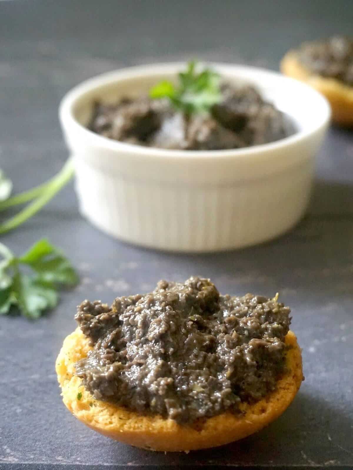 A slice of bread with tepenade and a white ramekin with more tapenade.