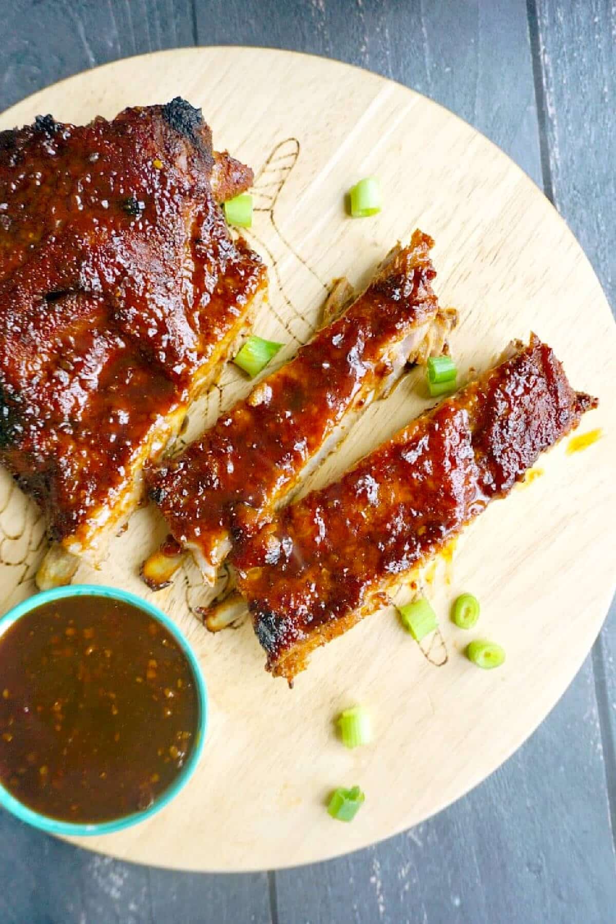 3 pork ribs on a wooden board with a small bowl of sauce.