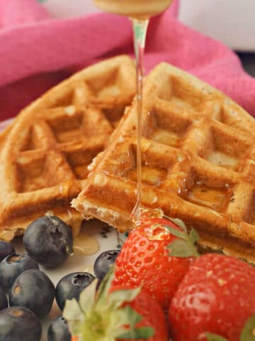 2 waffles on a plate with honey being drizzled over, blueberries and strawberries