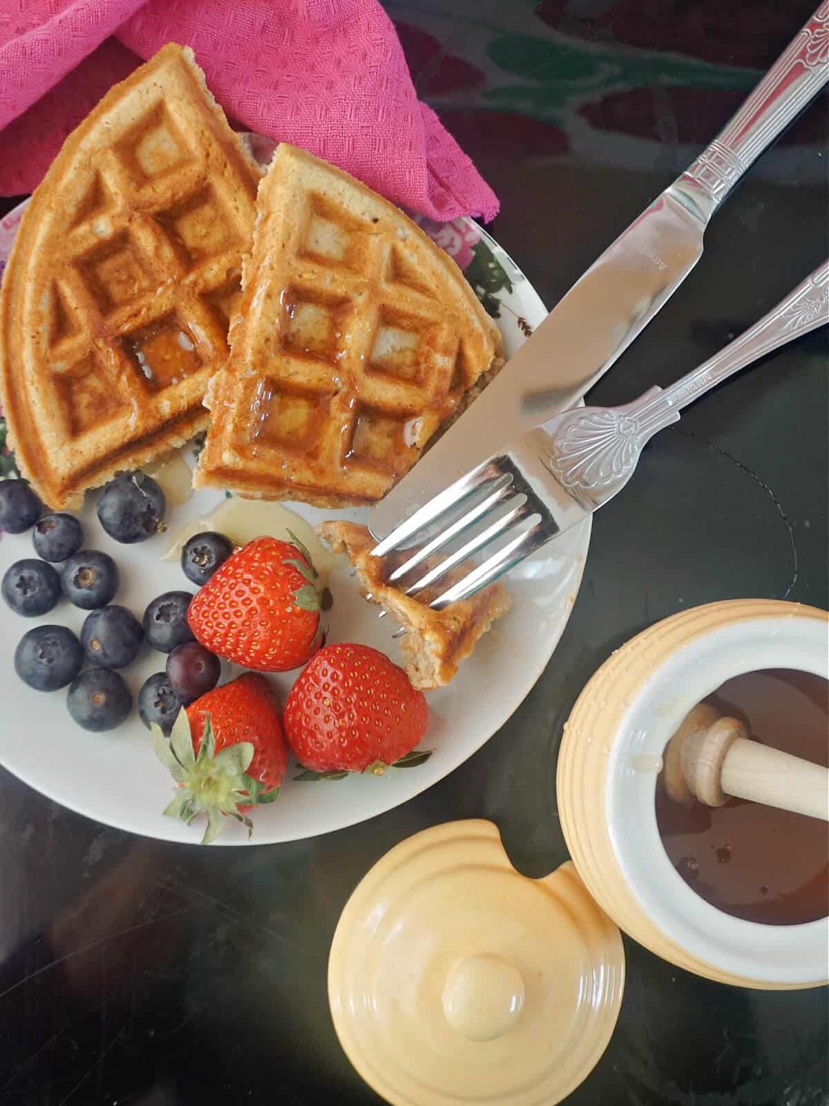 Overhead shoot of a plate with 2 waffles, blueberries and 3 strawberries, a pot of honey , a fork and a knife.