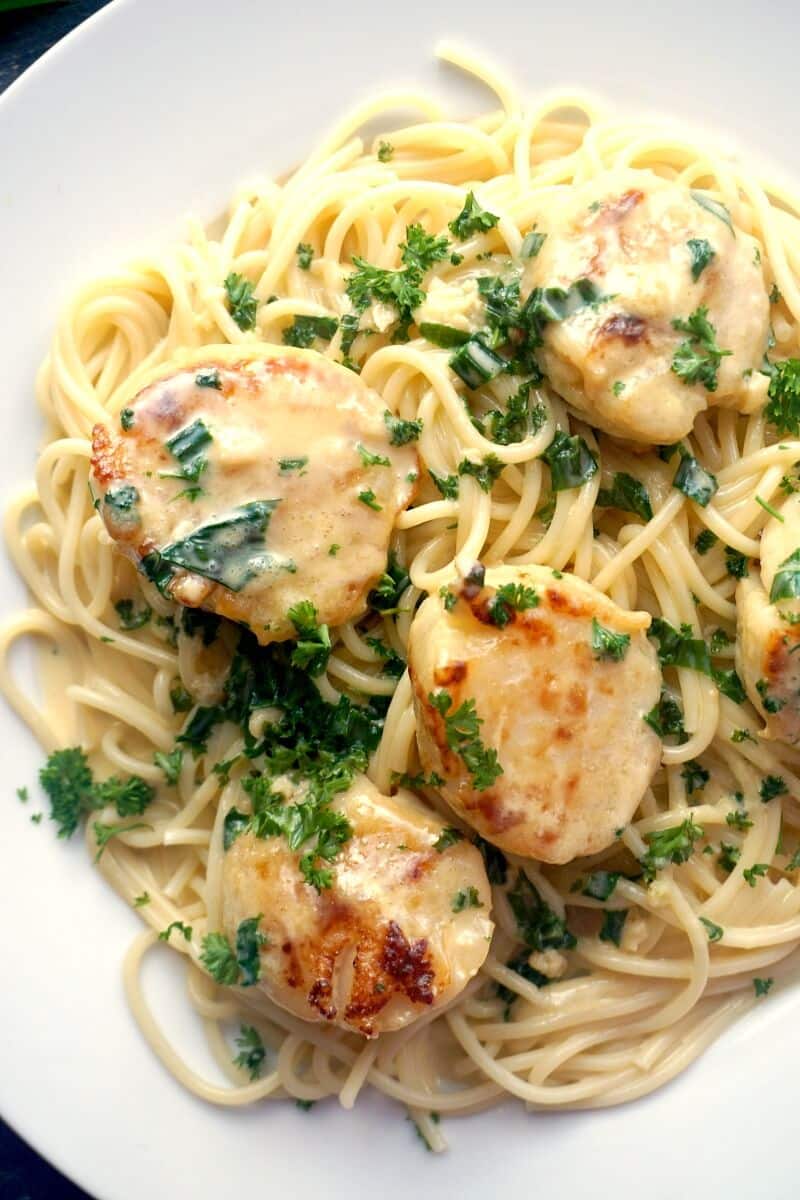 A white plate with spaghetti topped with seared scallops and a creamy herb sauce
