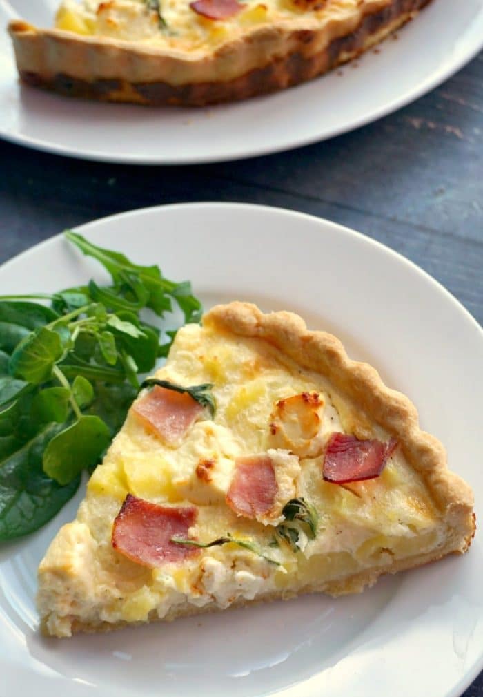 A white plate with a slice of potato and ham quiche and salad on the side