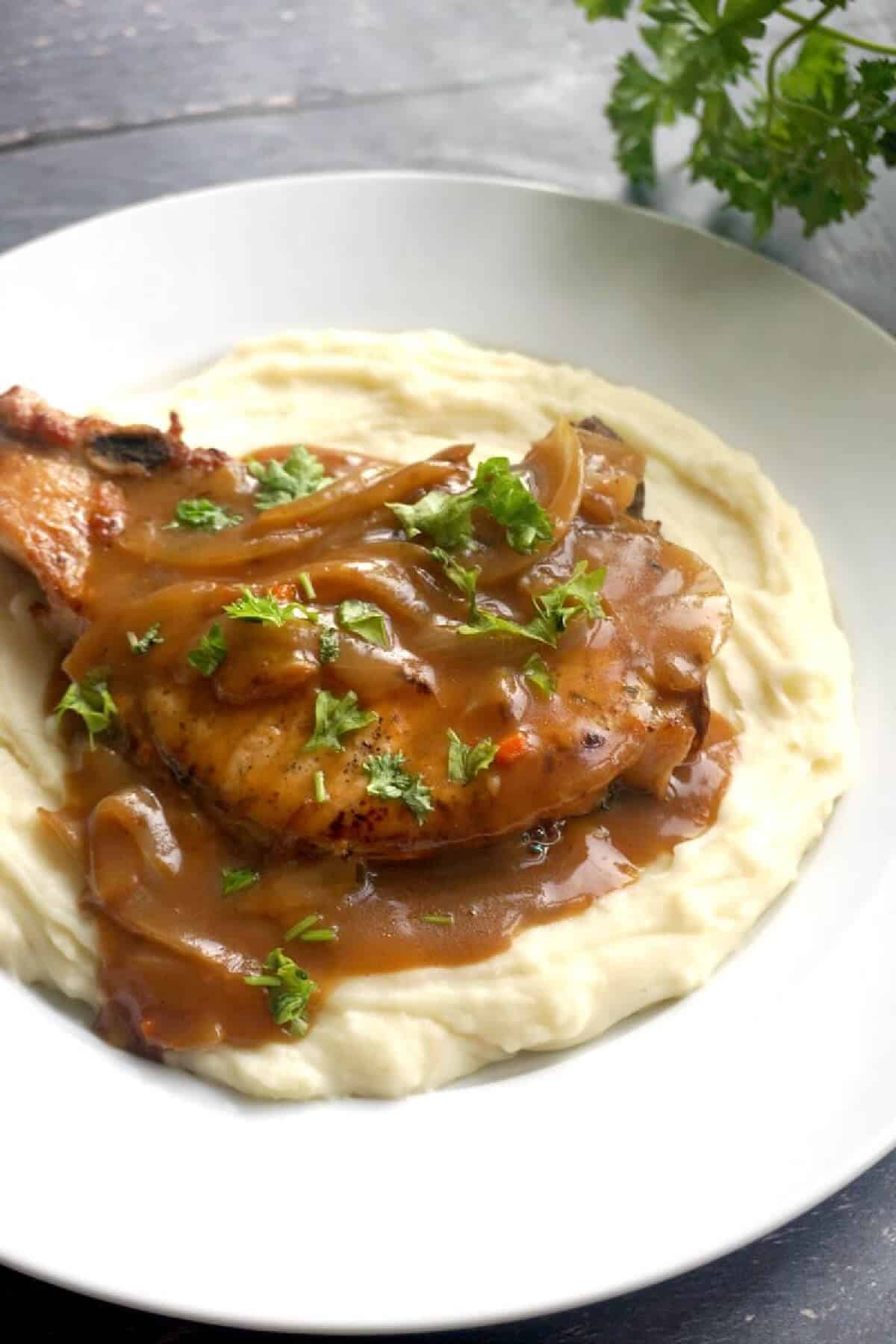 A white plate with mash topped with a pork chop in gravy.