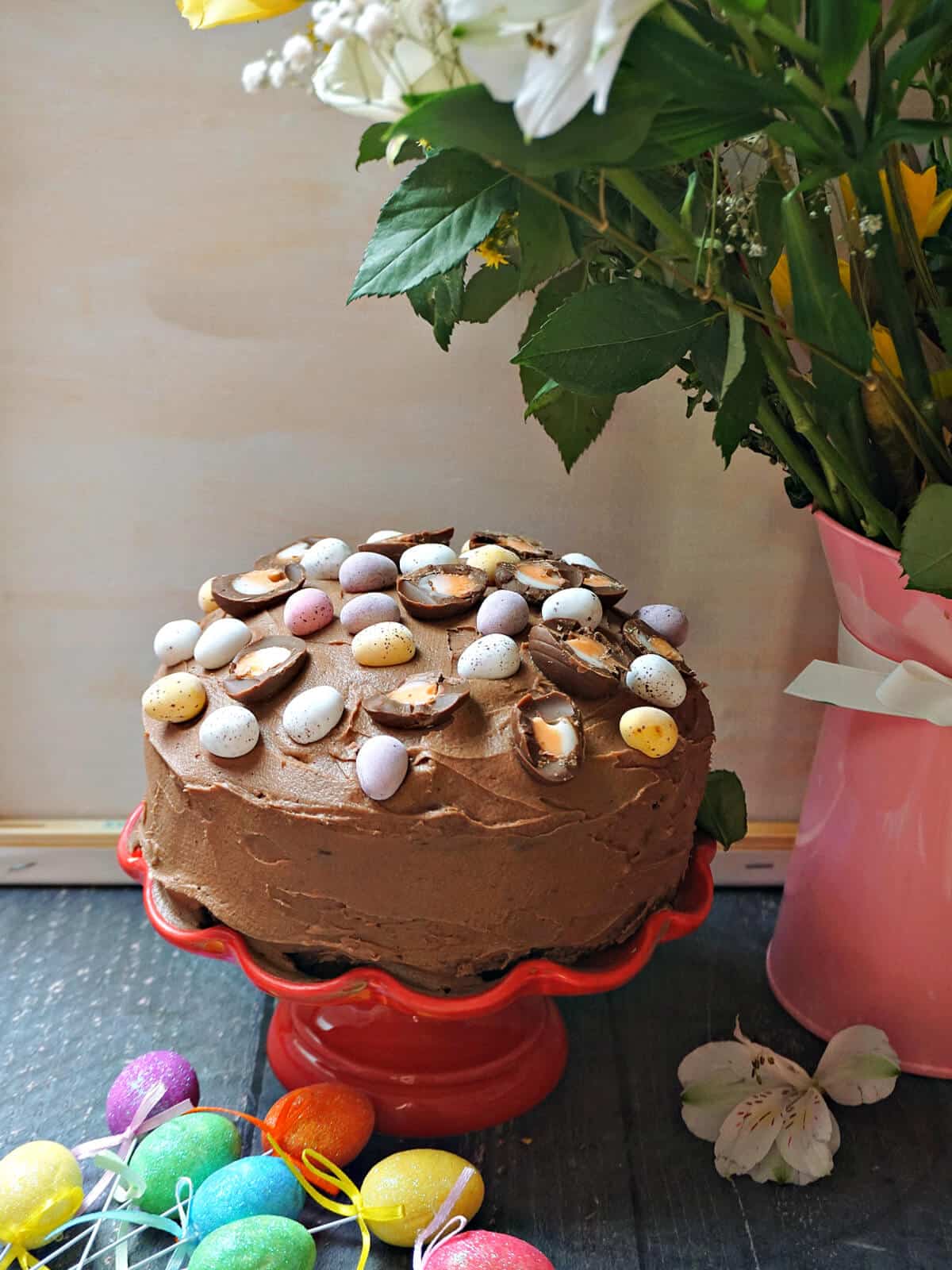 A chocolate cake decorated with Easter eggs on a red cake stand.