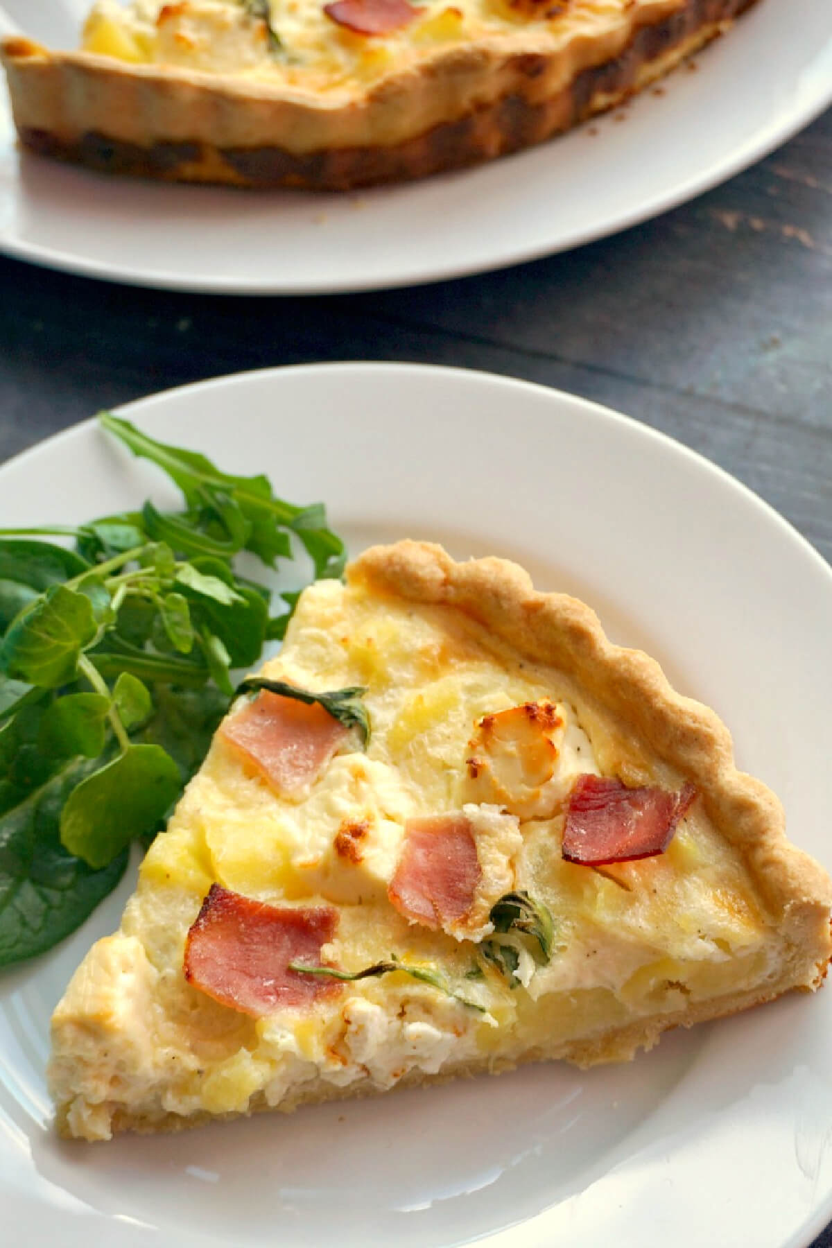 A white plate with a slice of ham and potato quiche with green salad on the side.
