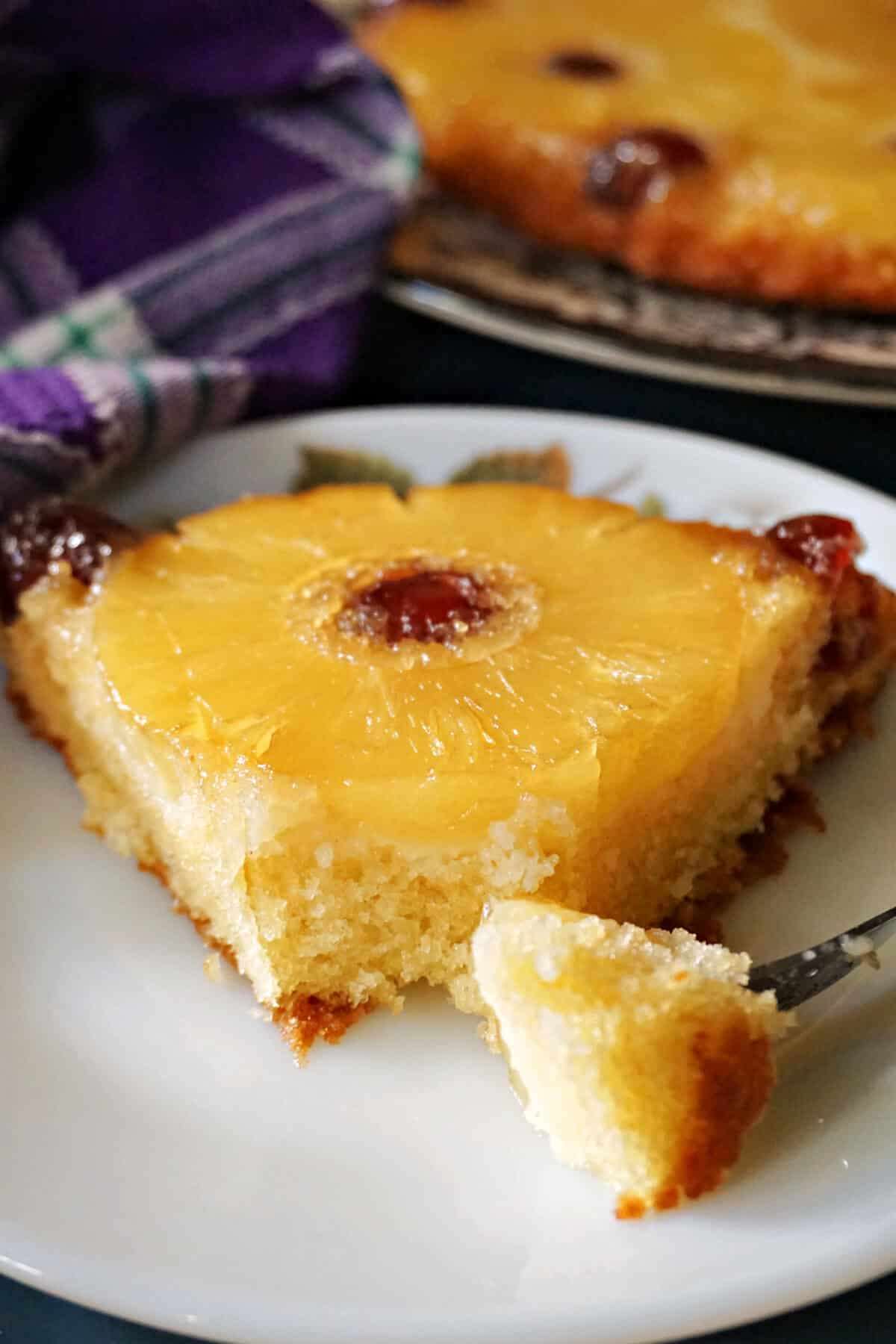 A white plate with a slice of pineapple upside down cake with a forkful of cake.