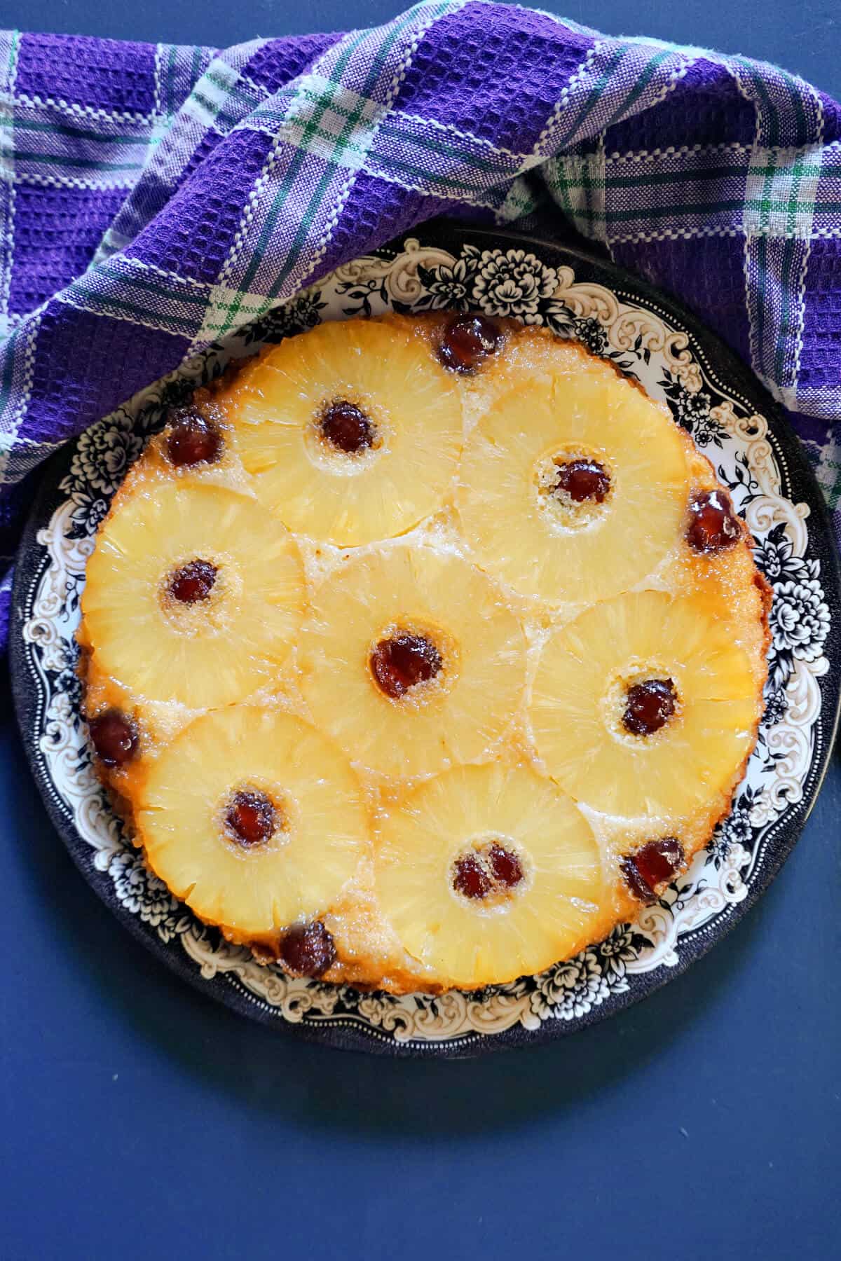 A plate with a pineapple upside-down cake and a purple tea towel around it.
