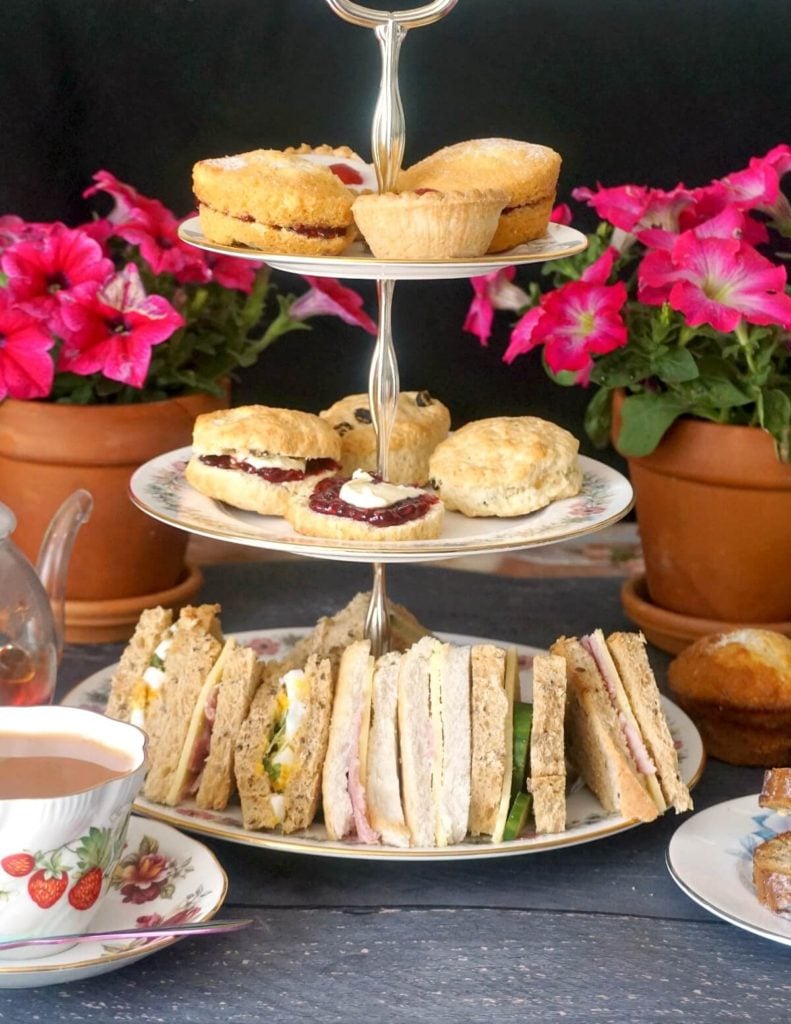 A cake stand with afternoon tea treats, a cup of tea and flowers in the background