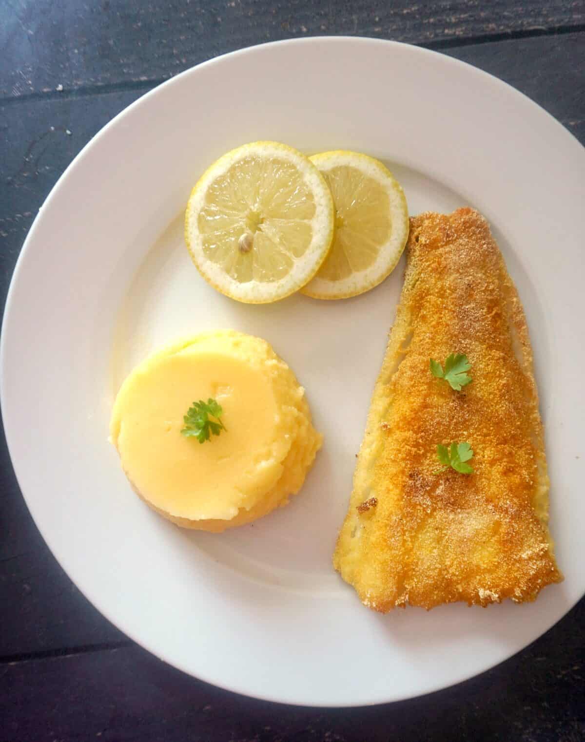 A white plate with a fried fish fillet, 2 slices of lemon and polenta.
