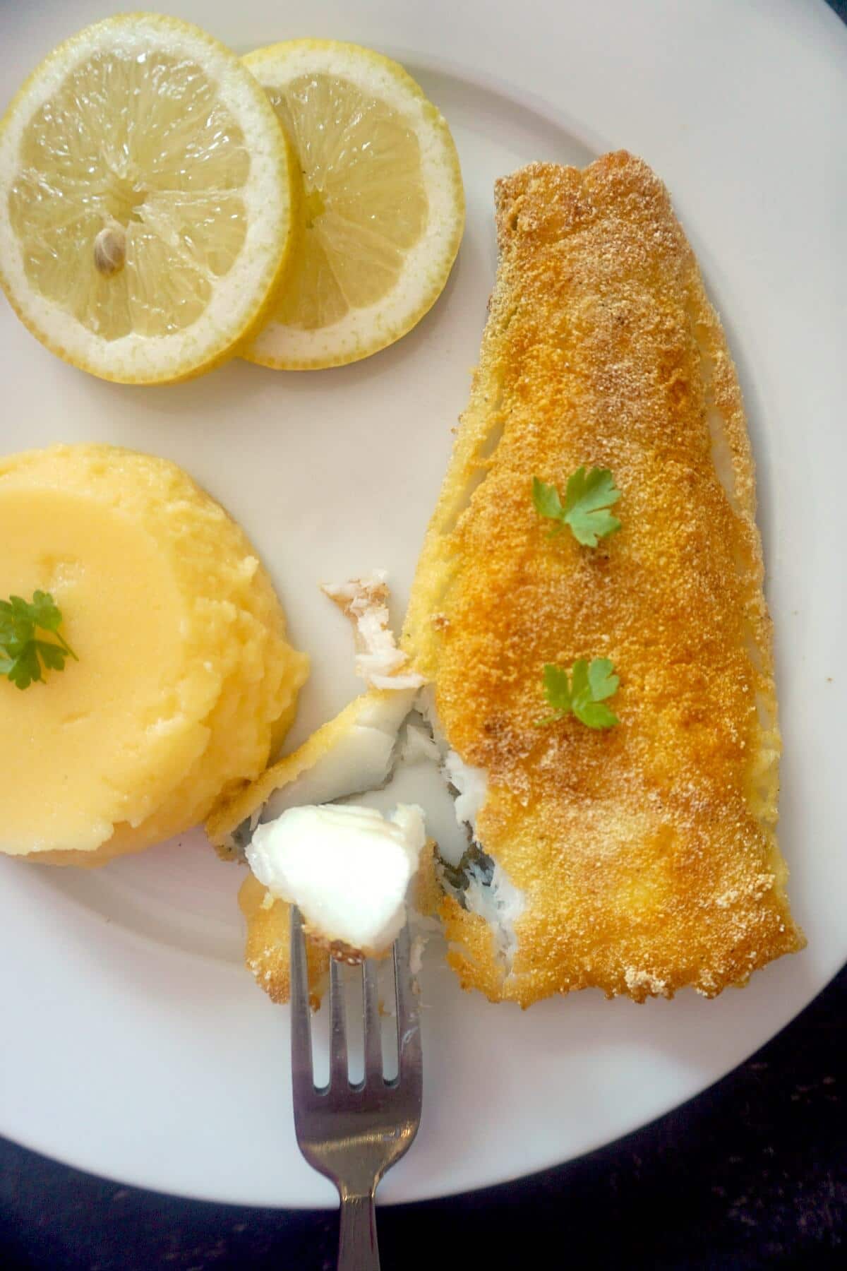 Overhead shoot of a cornmeal-crusted fried fish, 2 slices of lemon and polenta on a white plate.