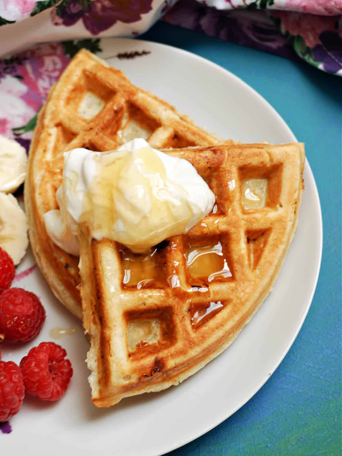 A white plate with 2 waffles topped with a dollop of yogurt and honey, banana slices and raspberries on the side.