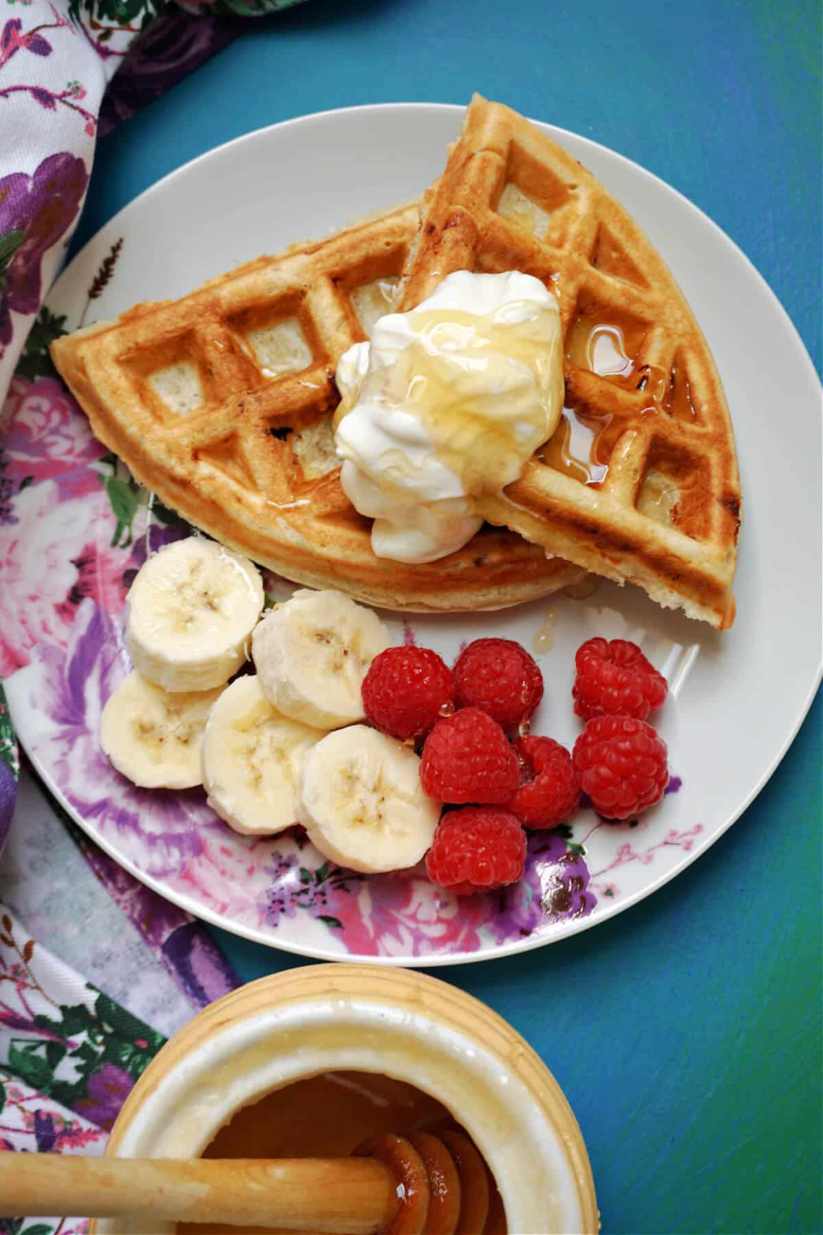 Overhead shoot of a white plate with 2 waffles topped with a dollop of yogurt and honey, plus banana slices and raspberries on the side.