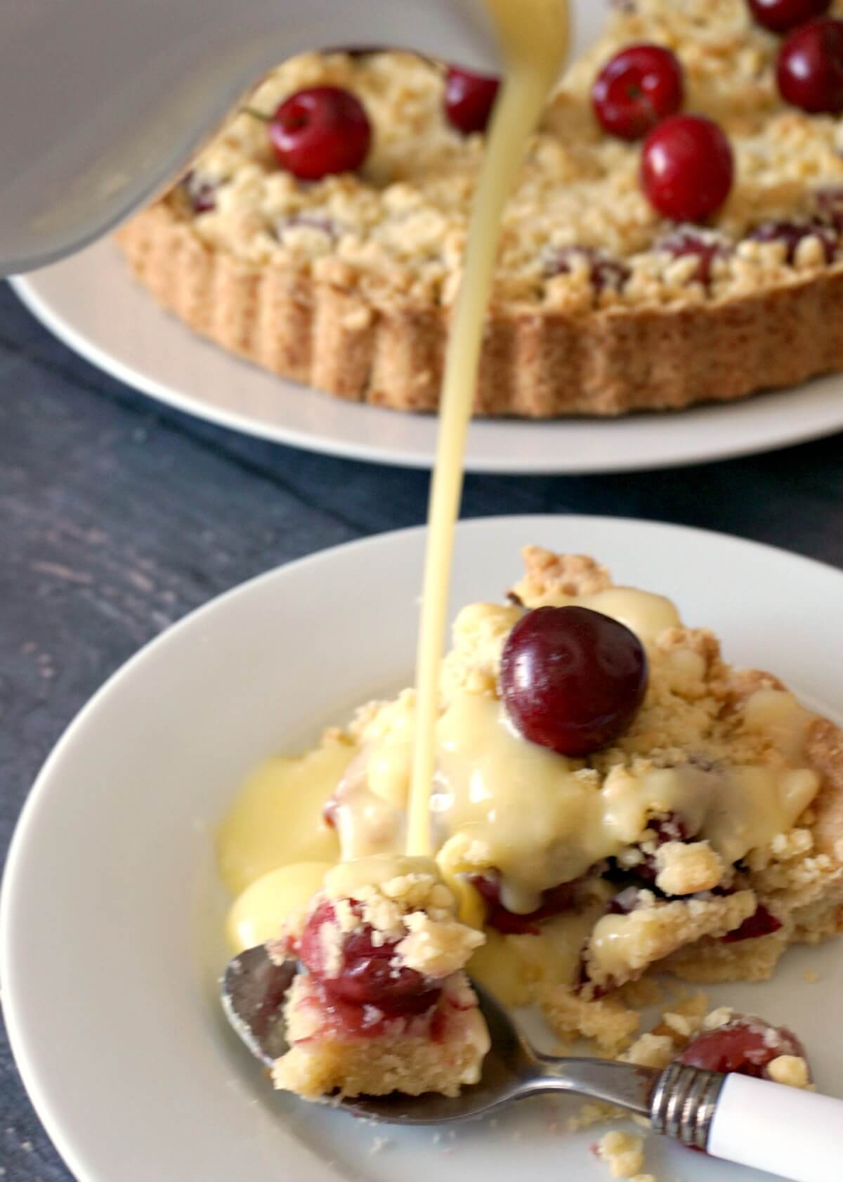 Custard being pour over a slice of cherry pie with more pie on a different plate in the background