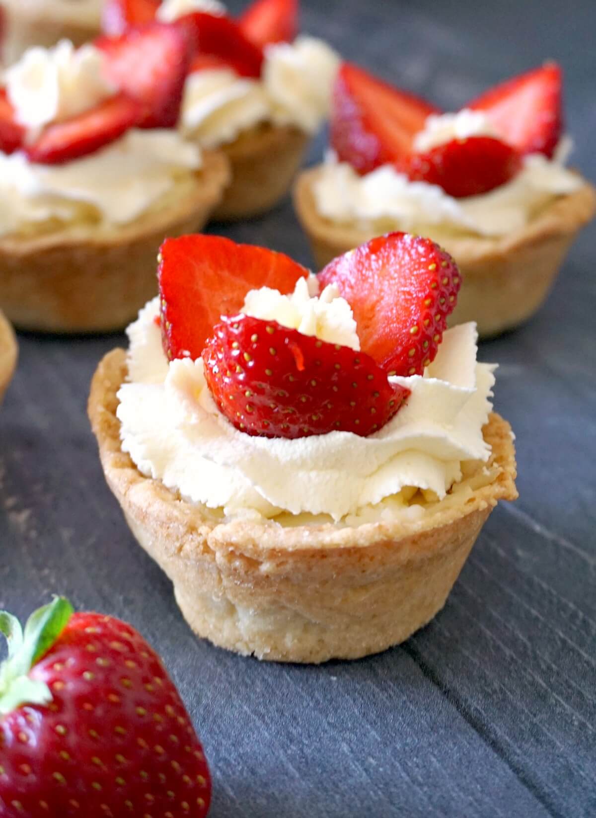 A tartlet with cream and 3 strawberry slices with other tartlets in the background