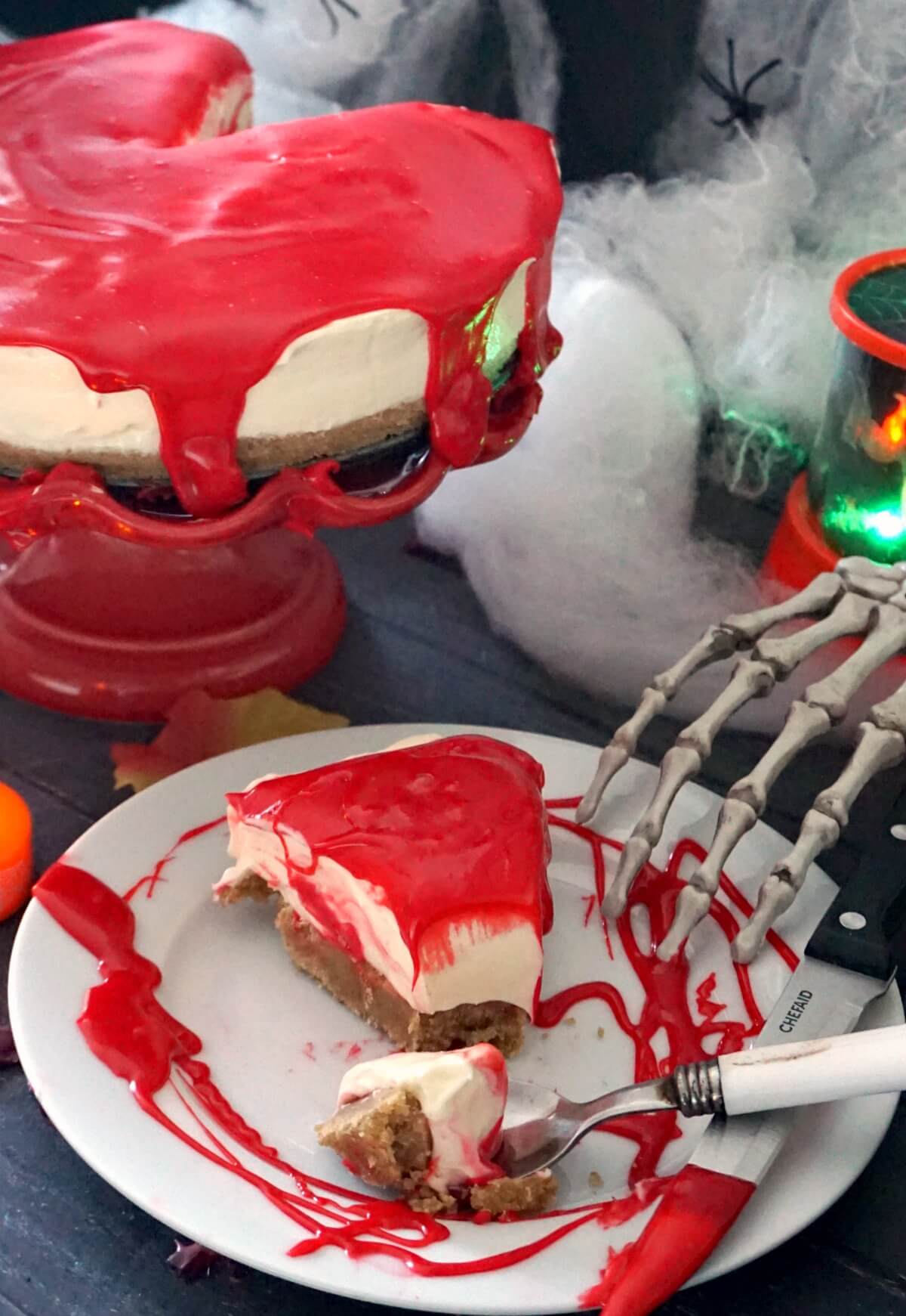 A slice of bloody cheesecake on a white plate with Halloween decorations around.