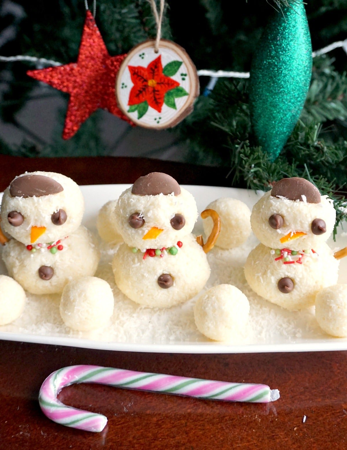 3 snowman truffles on a white rectangular plate with a Christmas tree in the background.