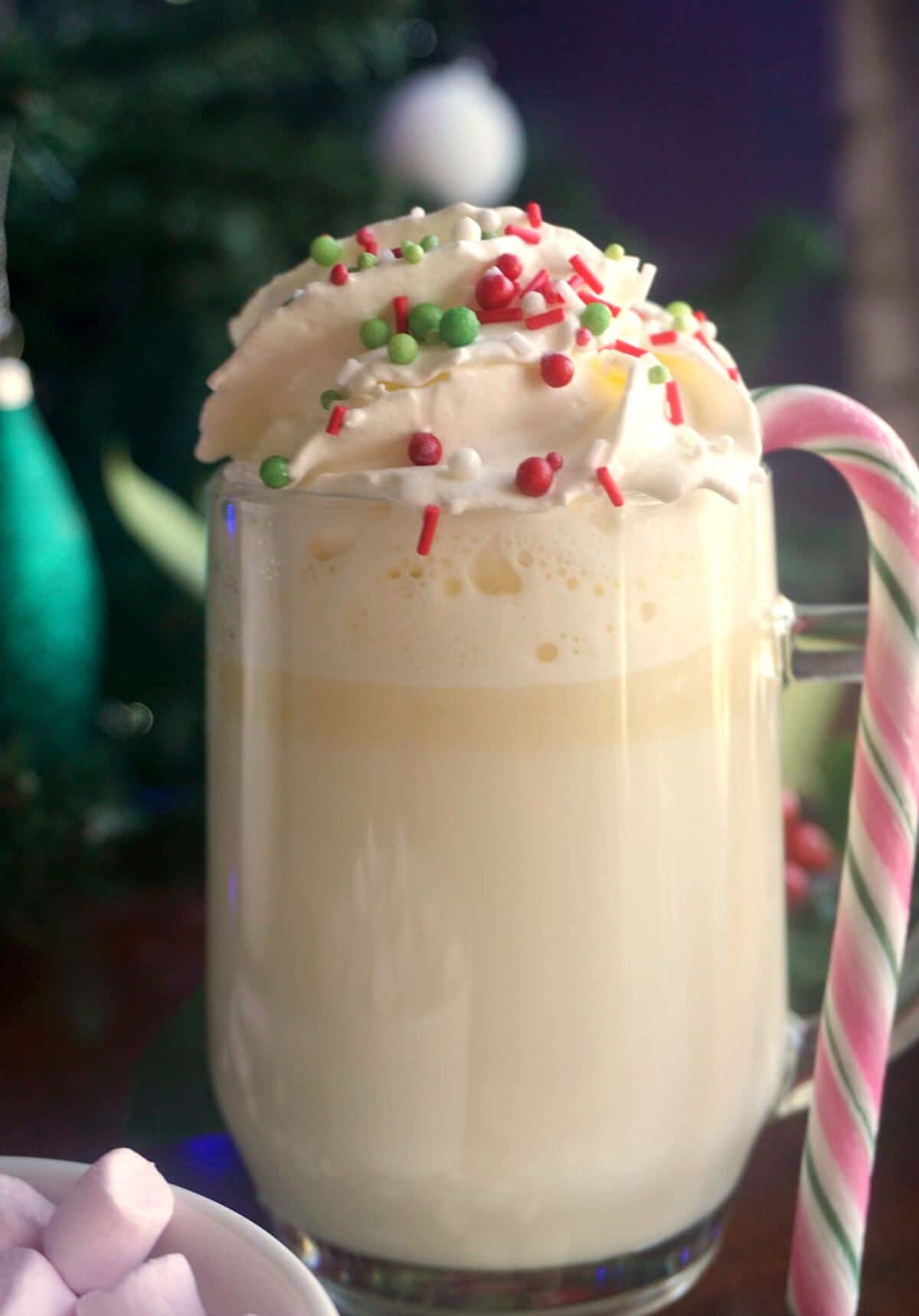 A glass of white hot chocolate with cream and sprinkles and a candy cane.