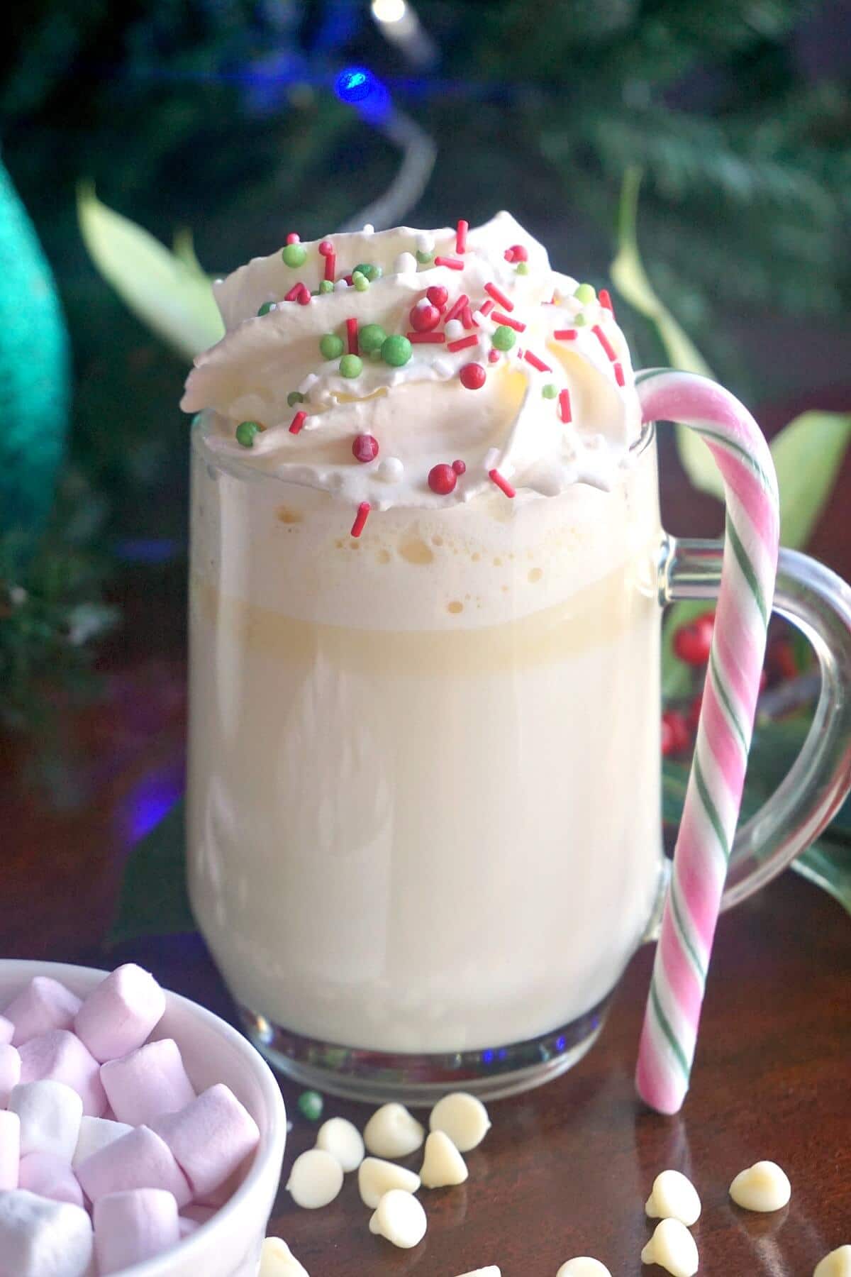 A glass of white hot chocolate with cream and sprinkles and a candy cane on the glass.