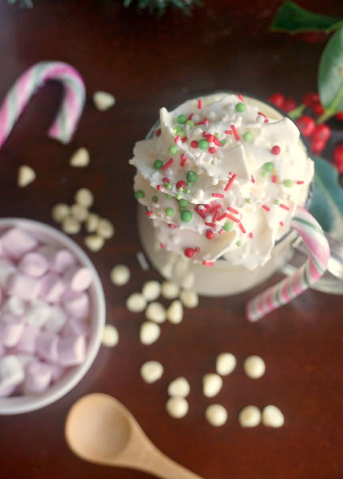 Overhead shot of a glass of white hot chocolate with cream and sprinkles, 2 candy canes and a ramekin of marshmallows.