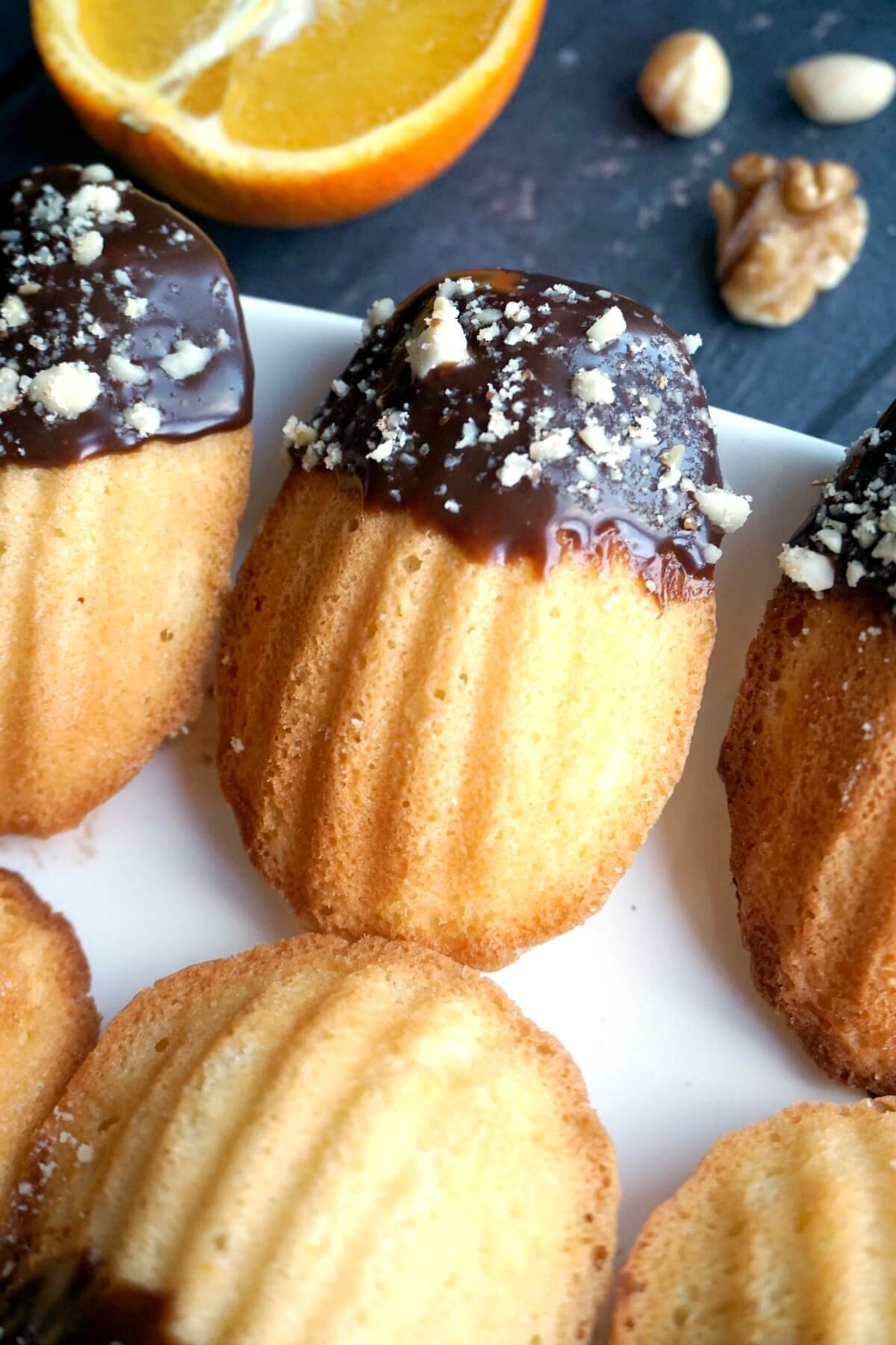Close-up shoot of orange madeleines dipped in chocolate and chopped nuts arranged on a white plate.