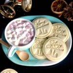 A blue plate with 4 Christmas stamped cookies and a mug of hot chocolate with marshmallows