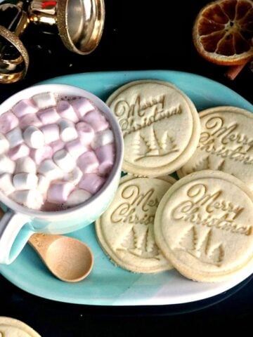 A blue plate with 4 Christmas stamped cookies and a mug of hot chocolate with marshmallows