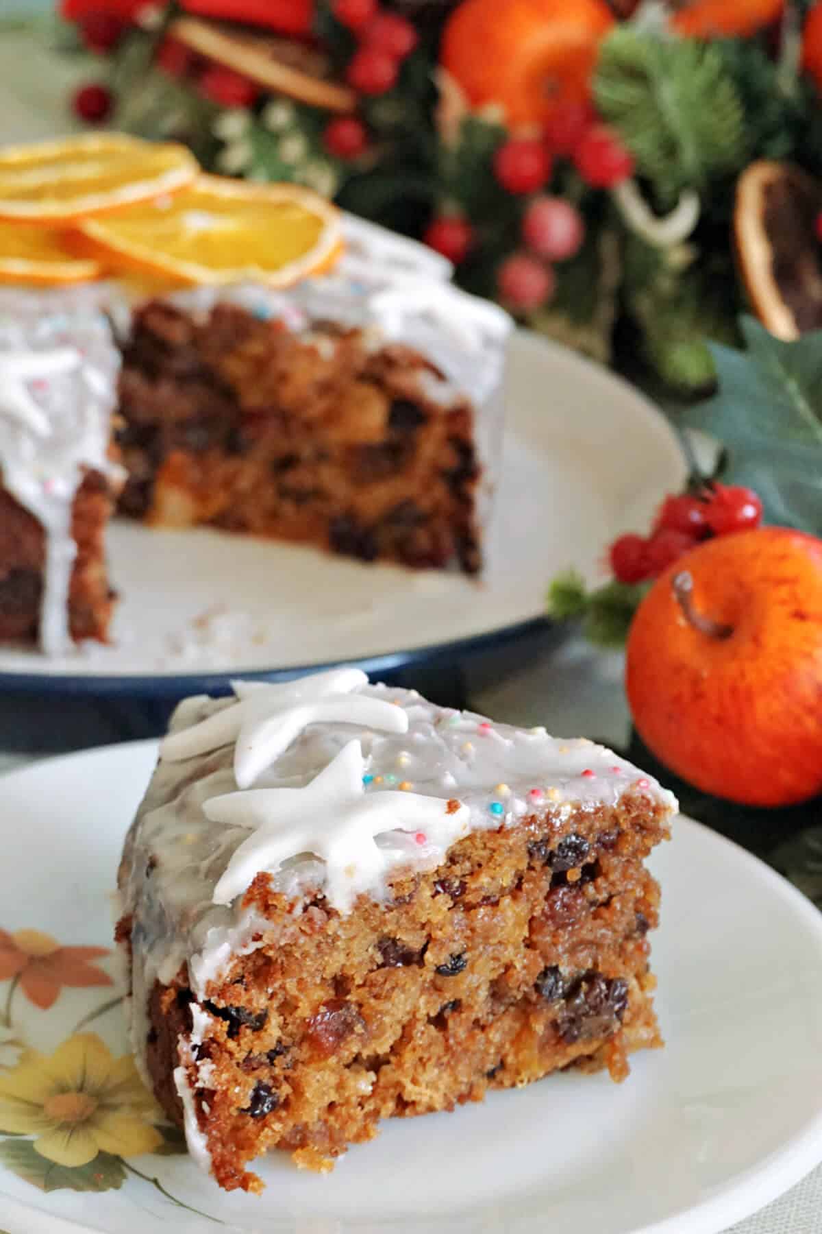 A white flowery plate with a slice of Christmas cake, a larger plate with more cake and Christmas decorations around.