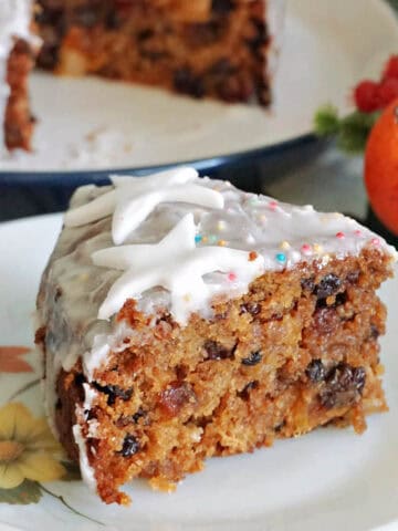 A slice of Christmas cake on a white flowery plate with a partially-visible larger plate with cake in the background.