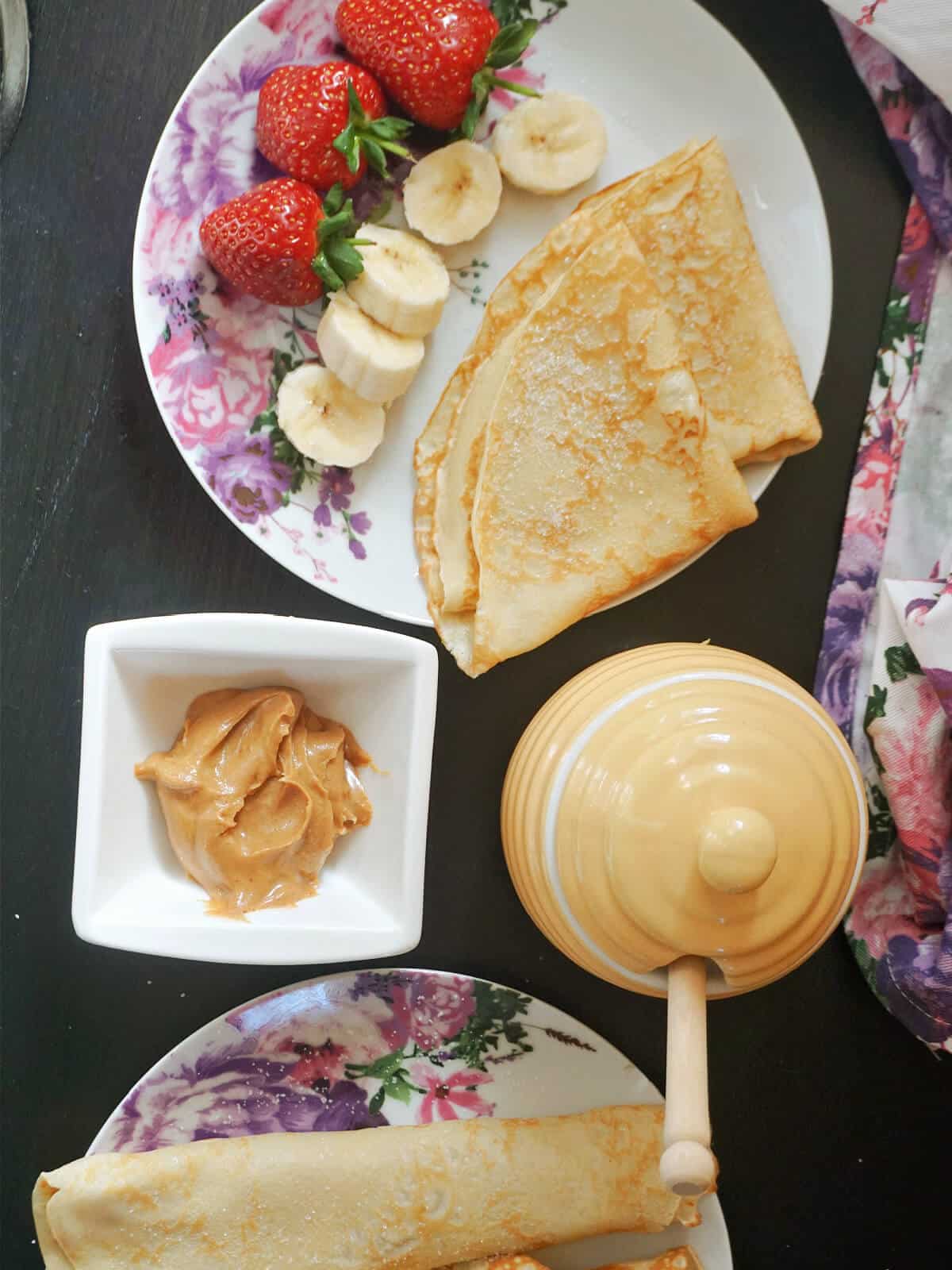 A white plate with 2 crepes, banana slices and strawberries, a ramekin with peanut butter, a honey oot and another plate with more crepes.