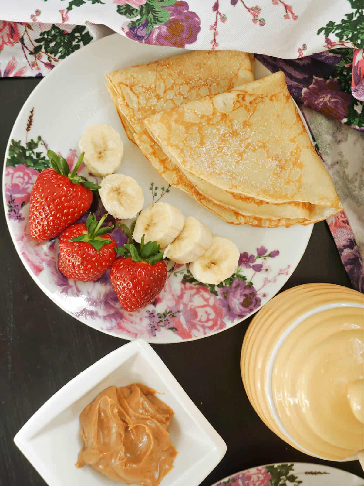 Overhead shoot of a plate with crepes, banana slices and strawberries, a ramekin with peanut butter and a pot of honey.