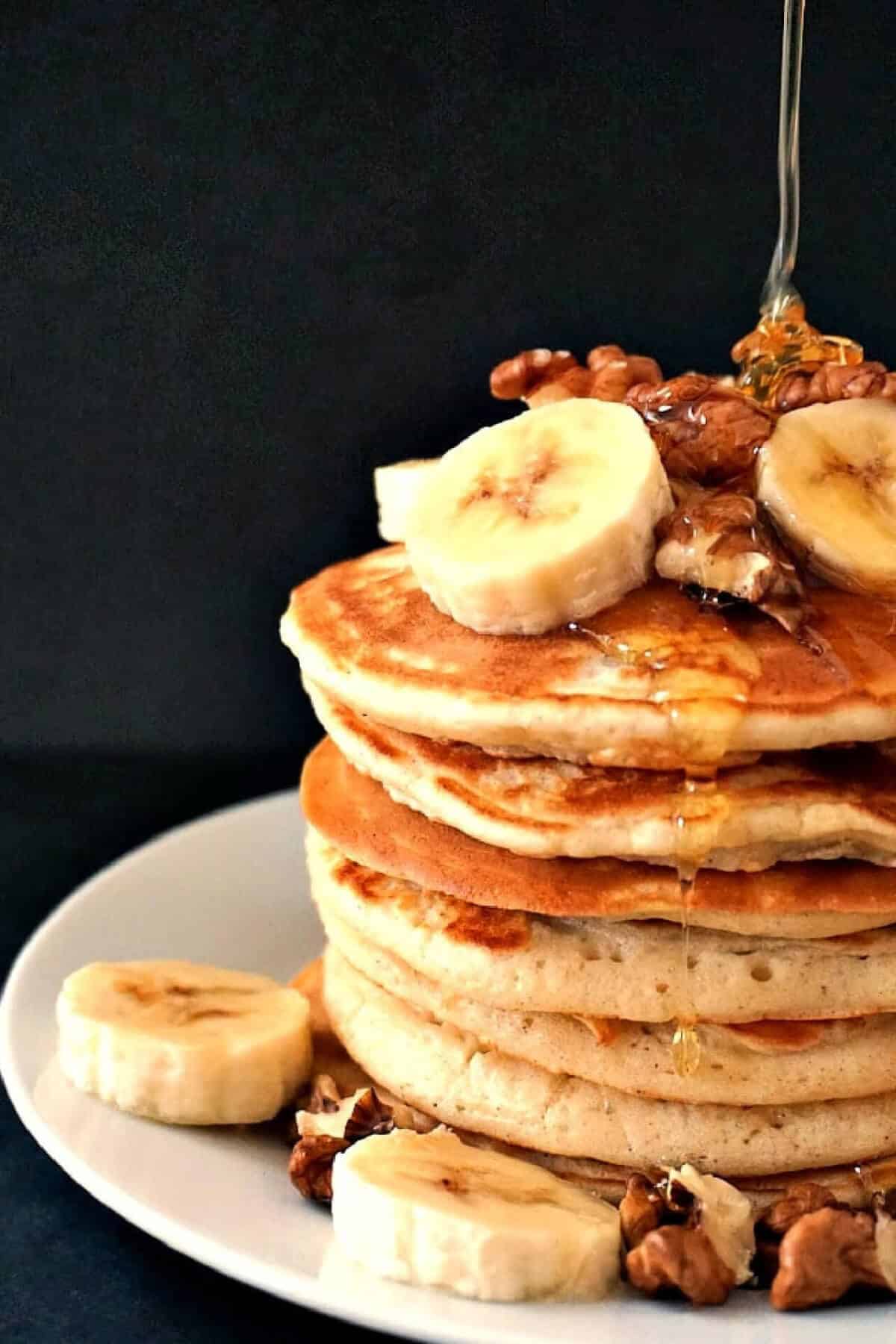 A pile of pancakes topped with walnuts and banana slices, and honey being squeezed over.