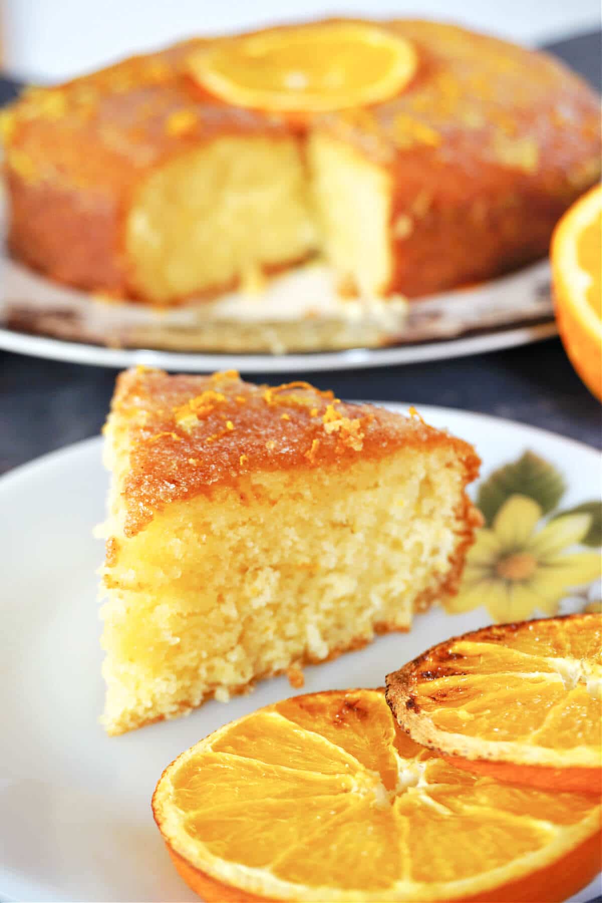 A slice of orange cake on a white plate with 2 slices of oranges and more cake in the background.