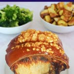 A roast pork with crackling on a white plate with 2 bowls of broccoli and roast potatoes in the background