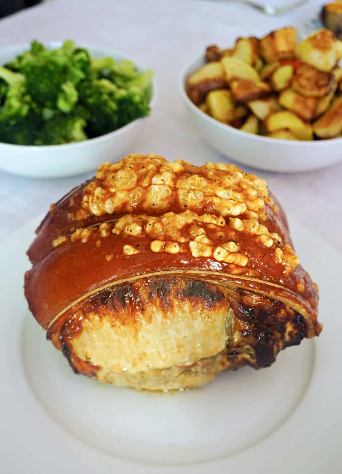 A roast pork joint on a white plate with broccoli and roasties behind
