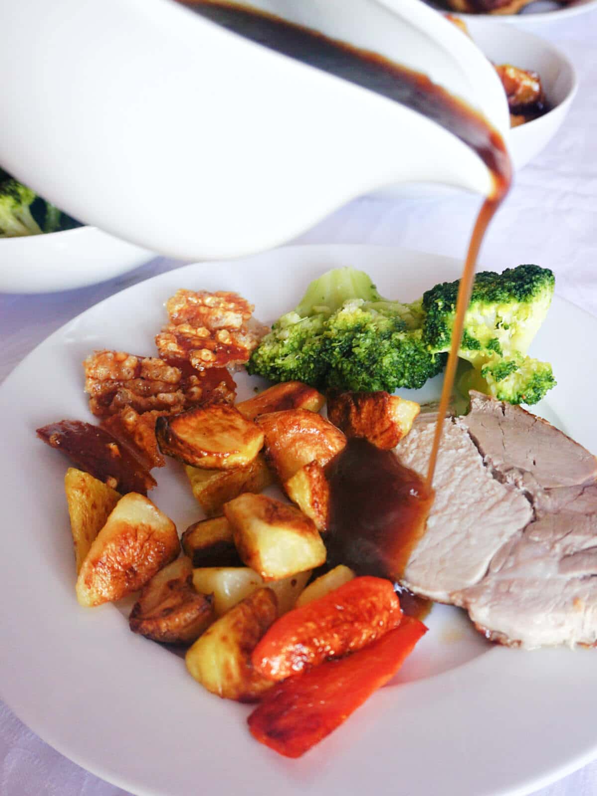 Gravy being poured over a slice of roast pork with vegetables on a white plate.