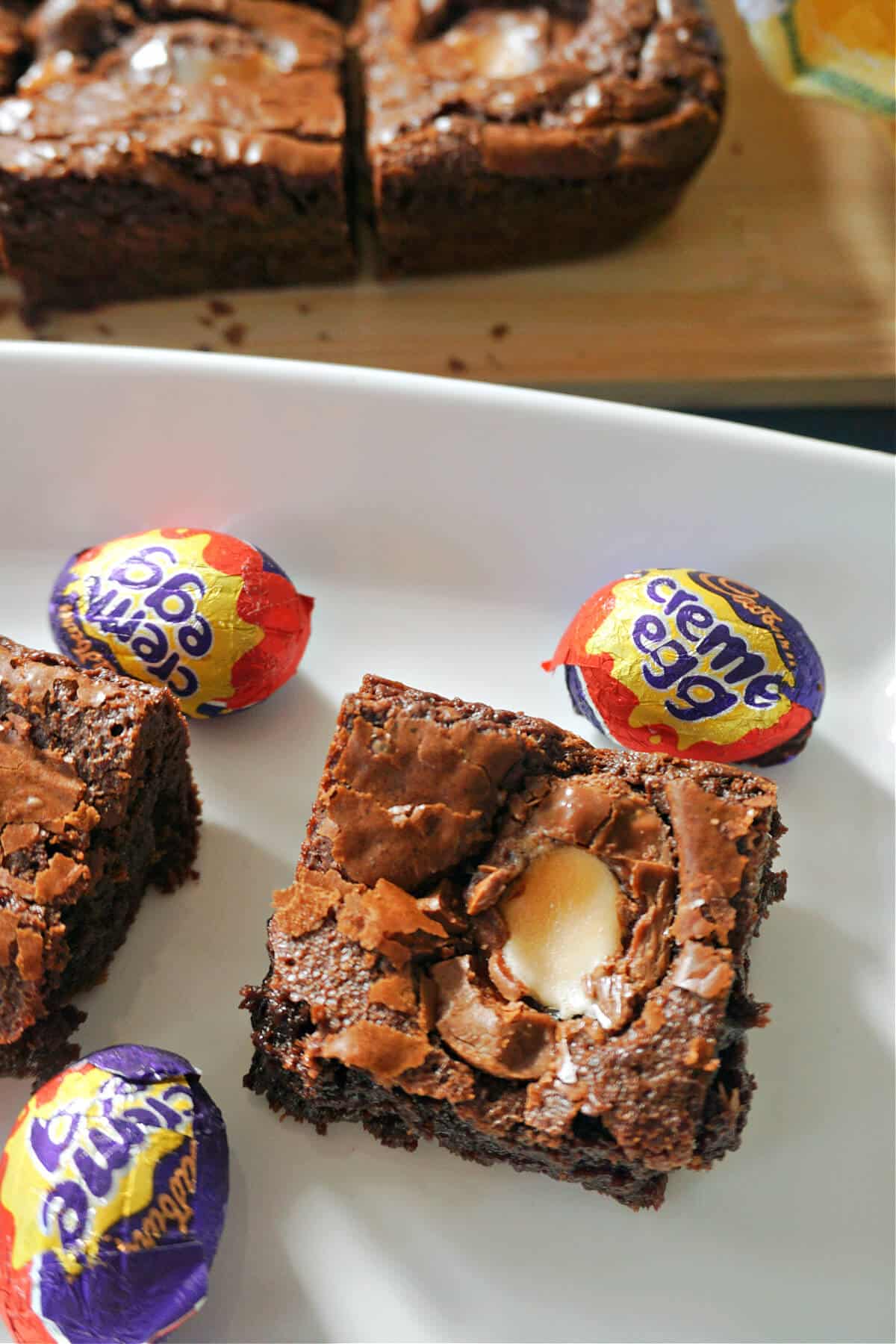 A slice of creme egg brownie on a white plate with chocolate eggs around.