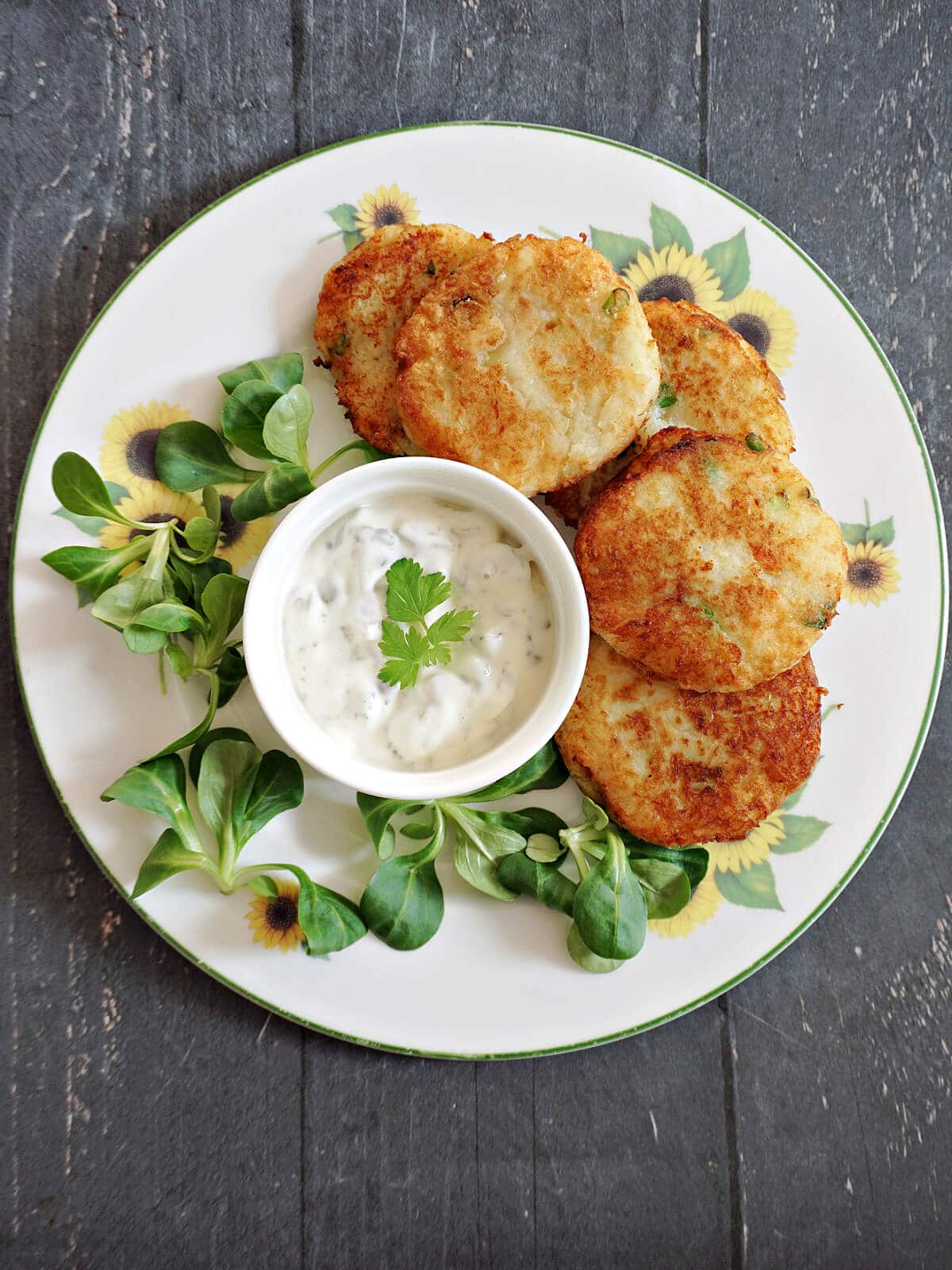 Overhead shot of a white plate with crab cakes, a ramekin of tartar sauce and green leaves around