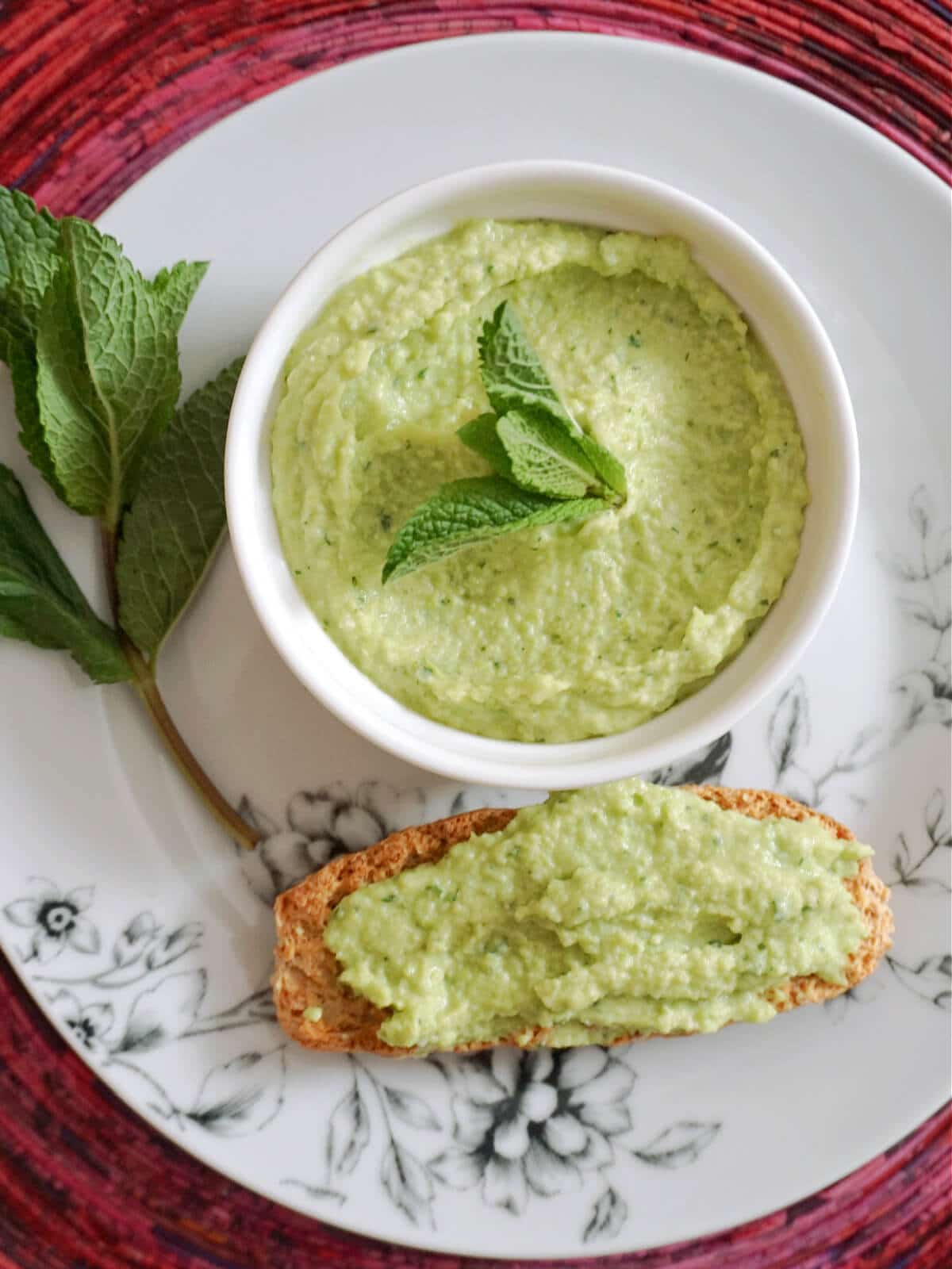 Overhead shot of a white plate with a ramekin of broad bean hummus, a slice of toast spread with dip and fresh mint leaves.