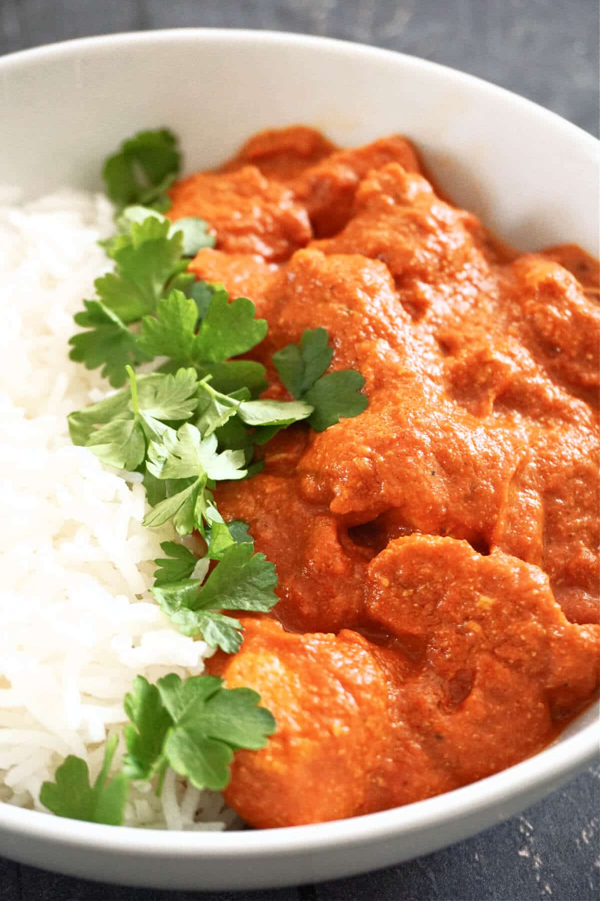 Close-up shot of a white bowl with chicken curry, rice and fresh parsley leaves