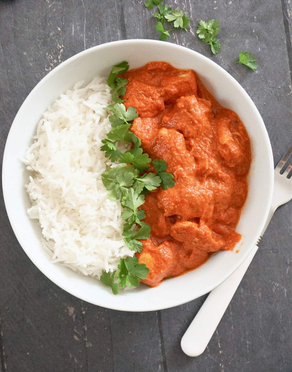 Overhead shot of a white bowl with butter chicken, basmati rice and fresh parsley leaves 