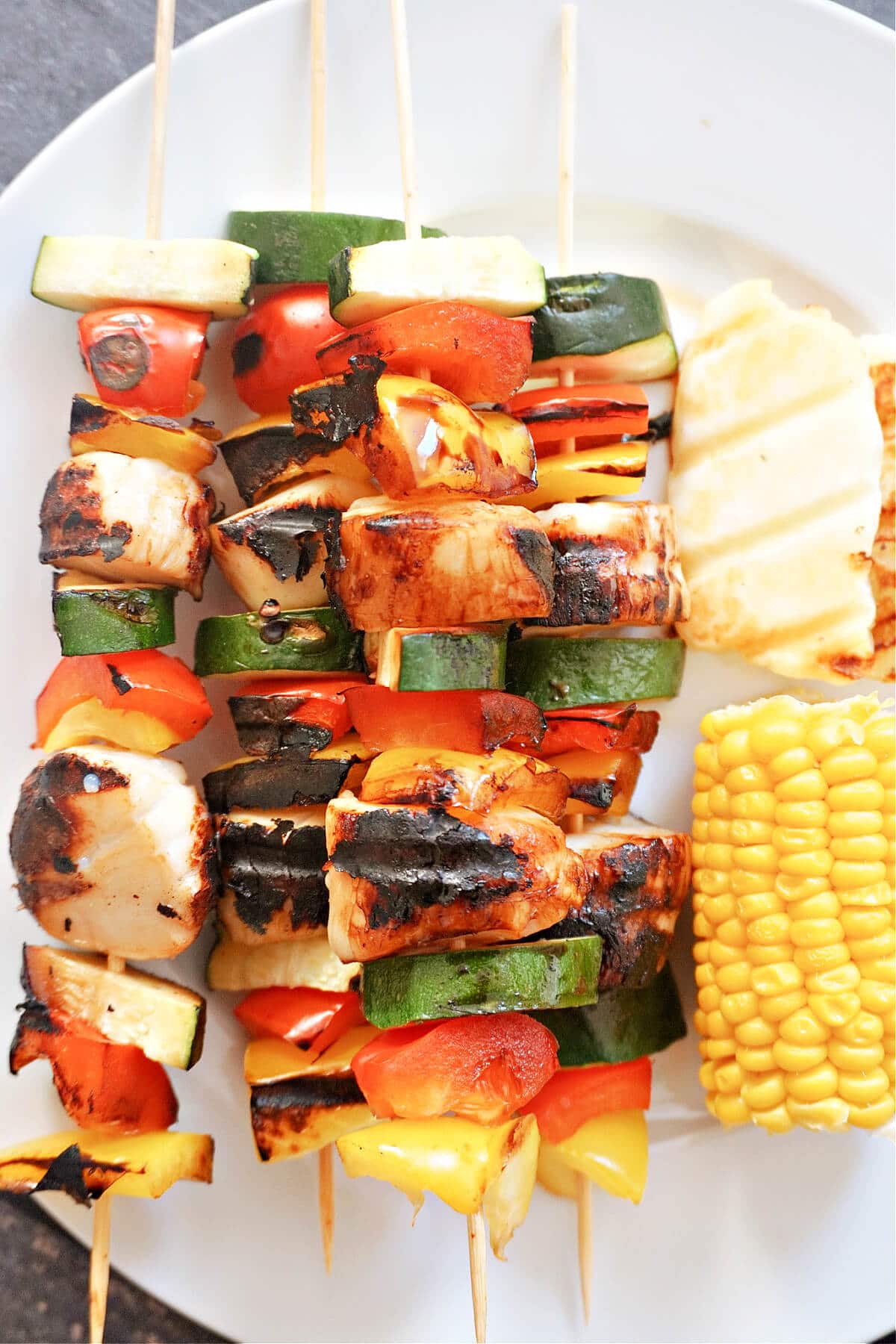 Close-up shot of 4 scallop and veggie skewers with corn on the cob and halloumi on a white plate.