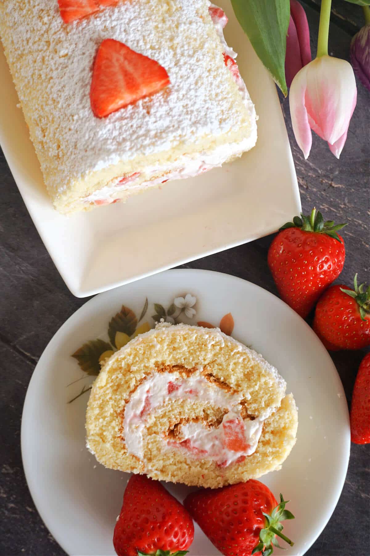 white plate with a slice of strawberry cake roll and another plate with the roll topped with strawberry slices.