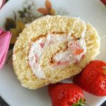 Overhead shot of a slice of strawberry cake roll on a white plate with 2 fresh strawberries on the side