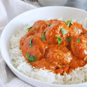 A white bowl with basmati rice topped with meatballs in curry sauce and fresh parsley