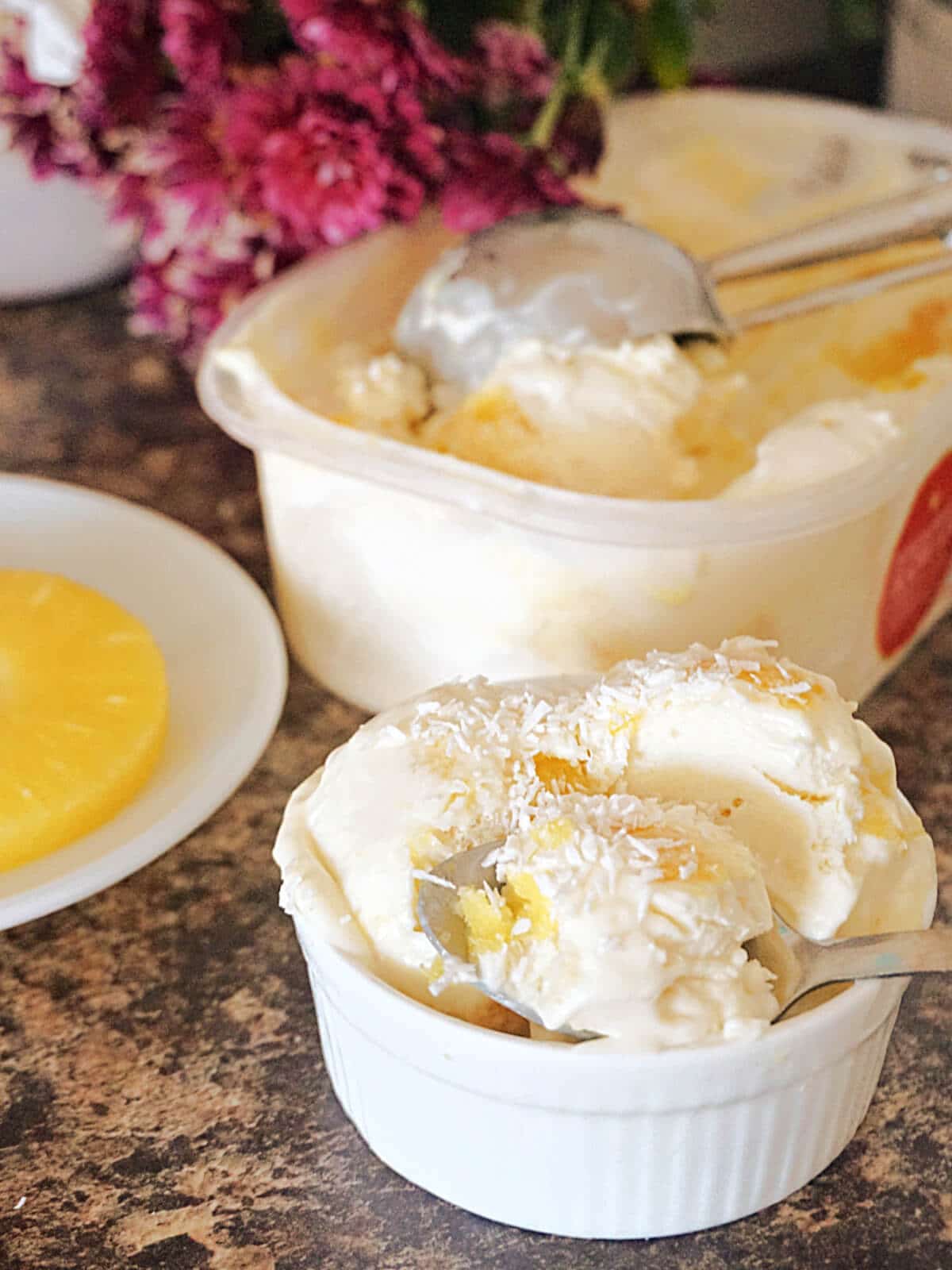 A white ramekin with 3 scoops of ice cream, a plate with a pineapple ring and a tub of more ice cream.