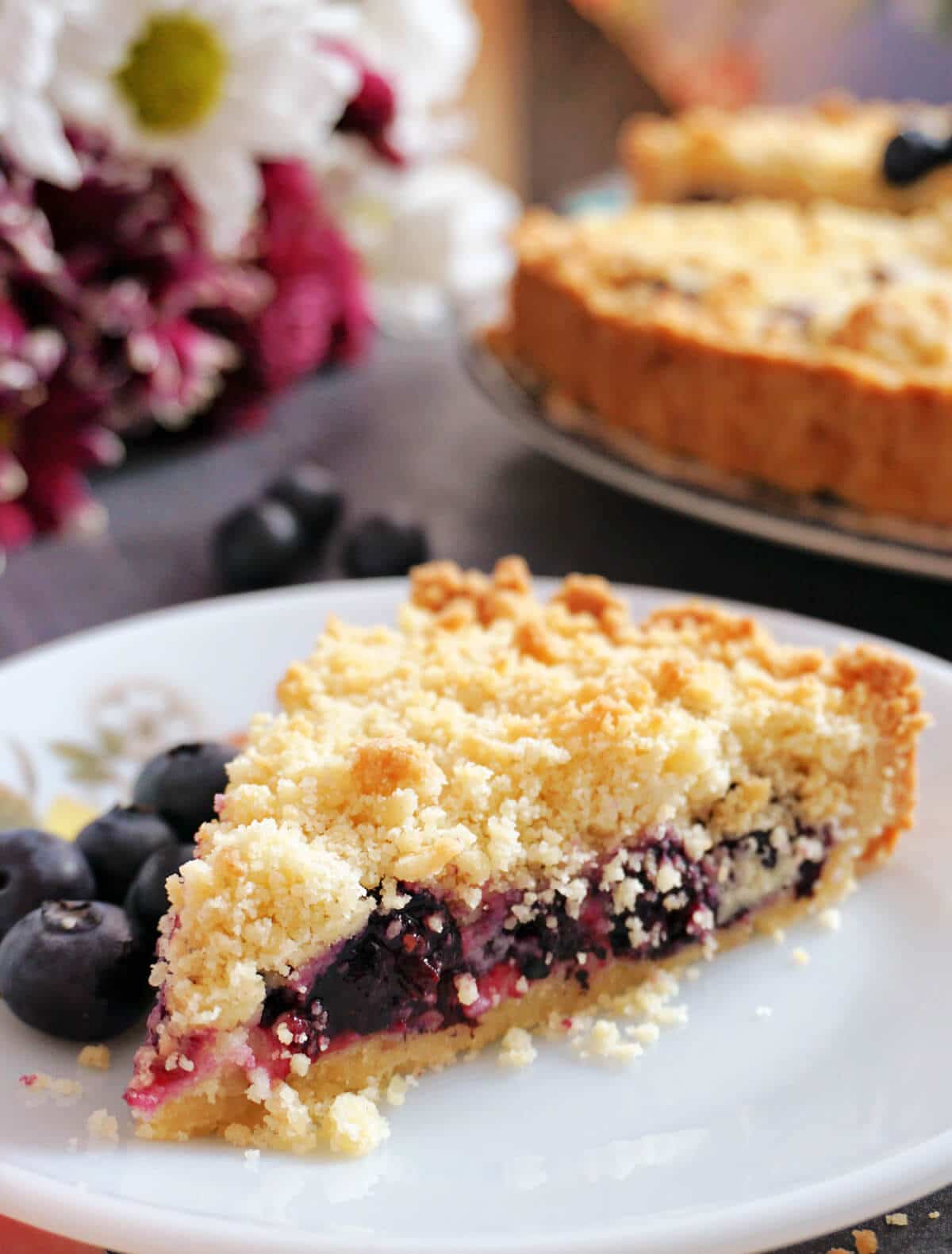A slice of blueberry pie on a white plate with blueberries around.