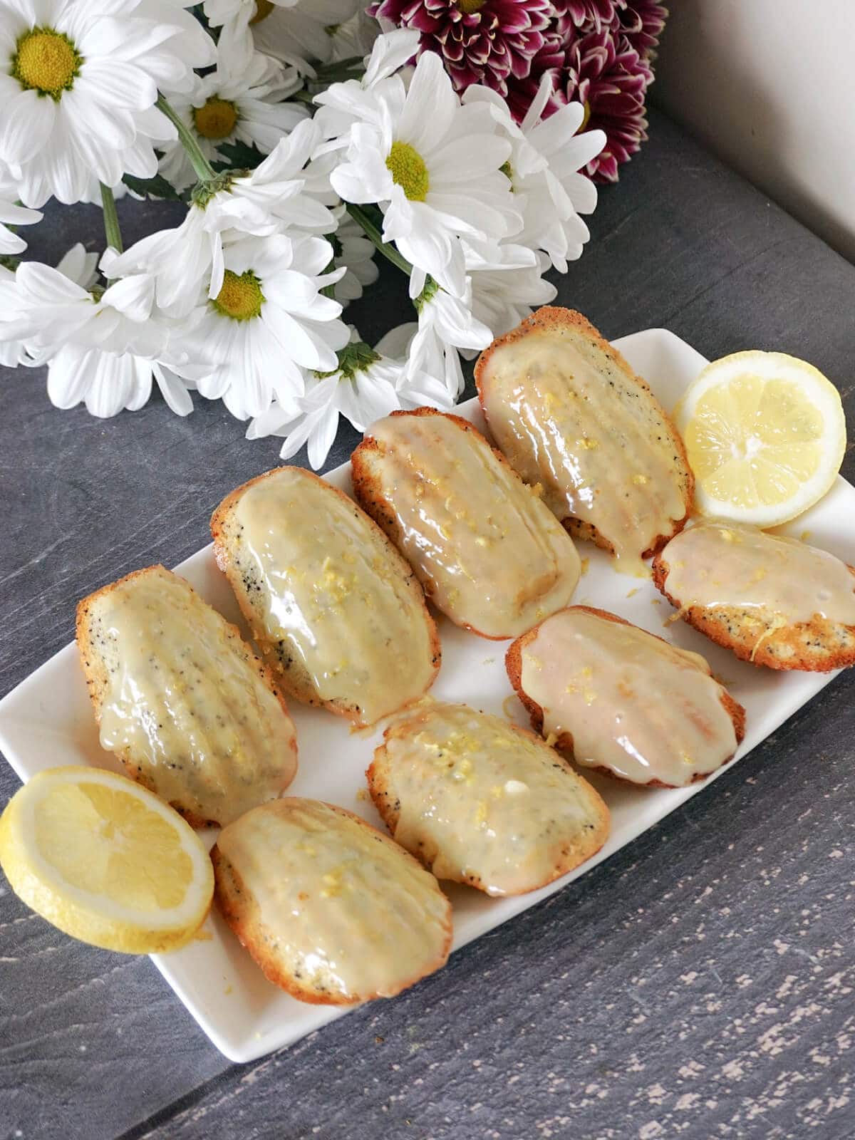 A white rectangle plate with 8 madeleines and 2 slices of lemon, with white flowers in the background