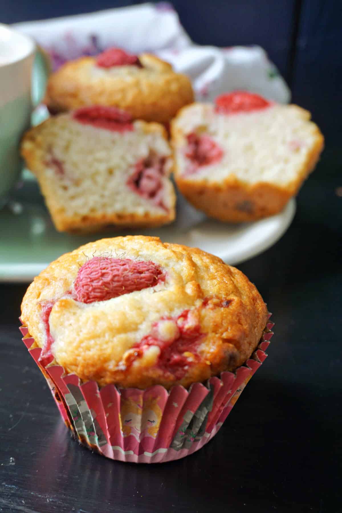 A raspberry muffin with a plate of 2 halves of a muffin and another whole muffin in the background.