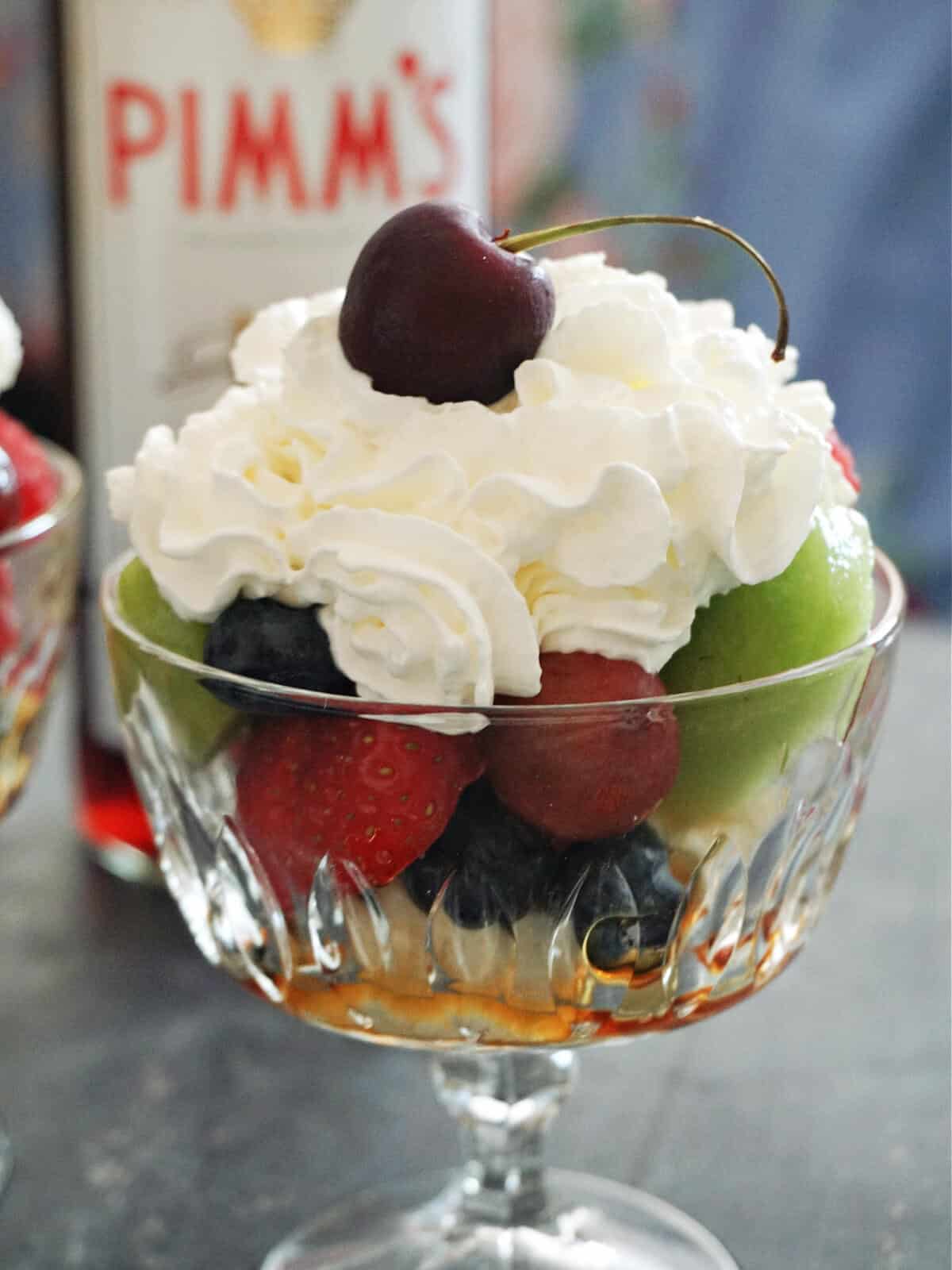 A glass of fruit salad topped with squirty cream and a cherry with a bottle of Pimm's in the background.