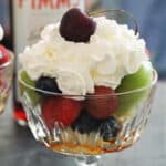 A glass with fruit salad topped with squirty cream and a bottle of pimms in the background
